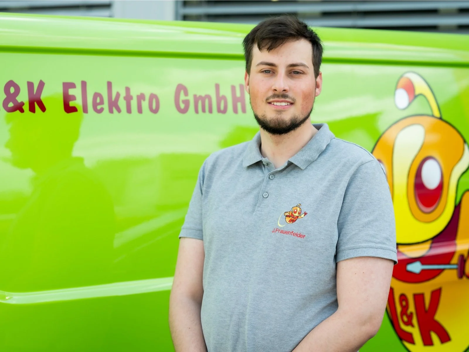 Ein junger Mann mit dunklem Haar und Bart steht vor einem grünen Fahrzeug mit Firmenlogo und der Beschriftung "&K Elektro GmbH". Er trägt ein graues Poloshirt mit einem kleinen Robotermotiv und dem Namen "J. Frauenfelder" darauf.