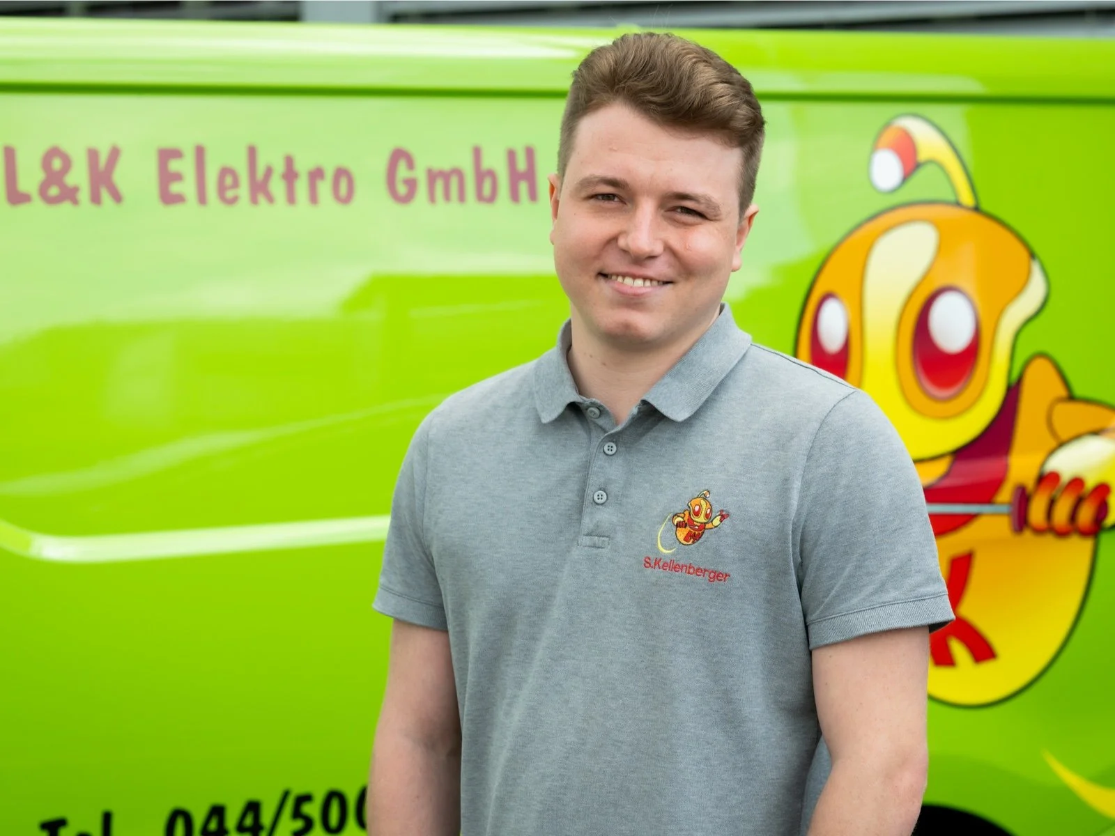 Junger Mann steht vor einem grünen Dienstfahrzeug mit einem Firmenlogo, trägt ein graues Polo-Shirt mit einem Cartoon-Branding und dem Namen S. Kellenberger.
