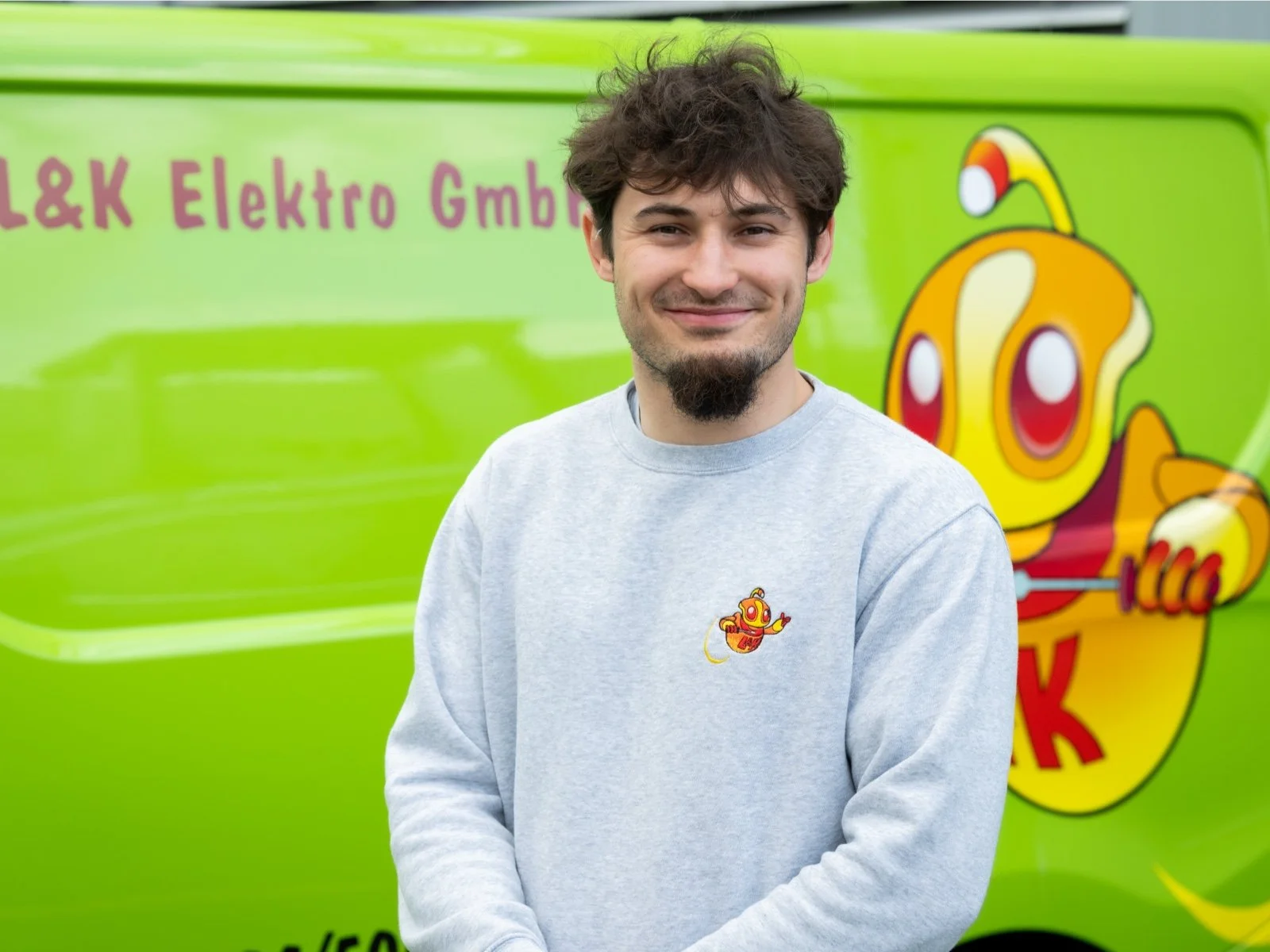 Junge Mann steht vor einem grünen Lieferwagen mit einer Cartoon-Charakter-Illustration und dem Text 'L&K Elektro GmbH'. Er trägt ein graues Sweatshirt mit einem kleinen Cartoon-Charakter auf der Brust. Er lächelt in die Kamera.