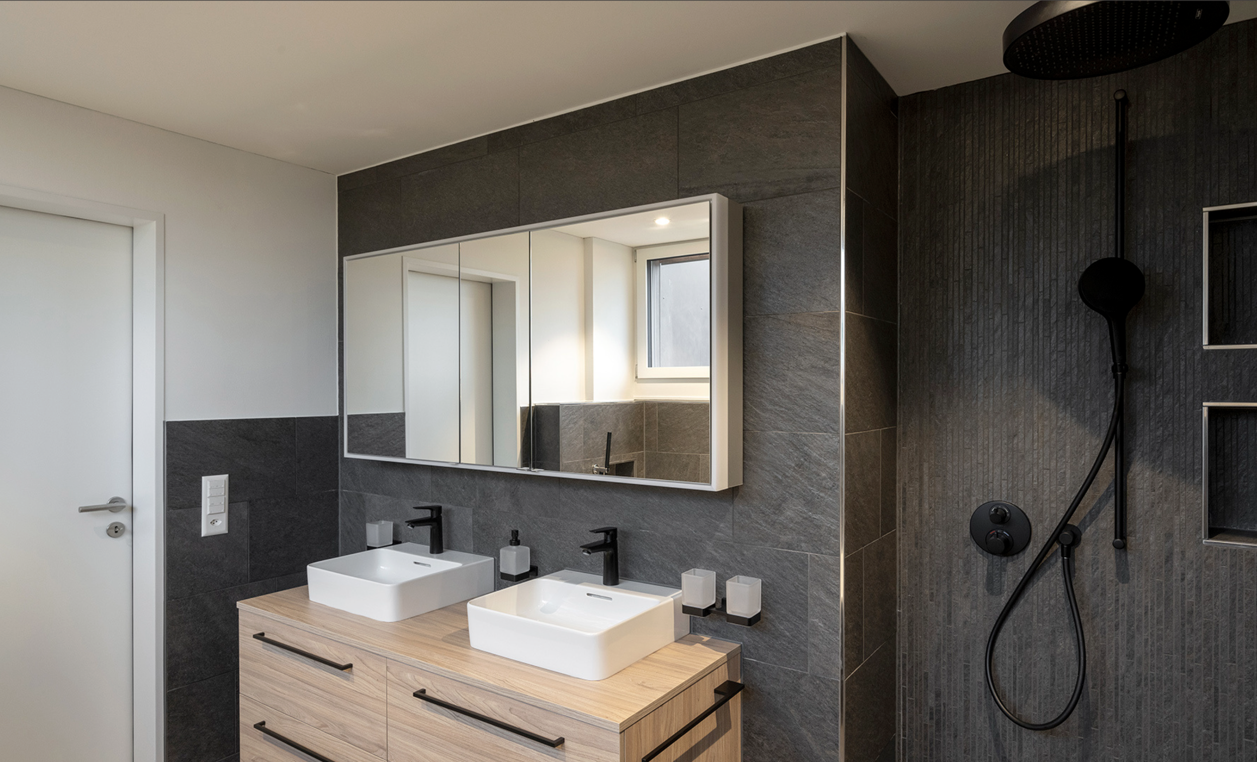 Moderner Badezimmer mit Doppel-Waschbecken, großen Spiegel, dunklen Wandfliesen, Holzunterschrank, schwarzer Armatur, Dusche mit schwarzem Duschkopf und HDTV