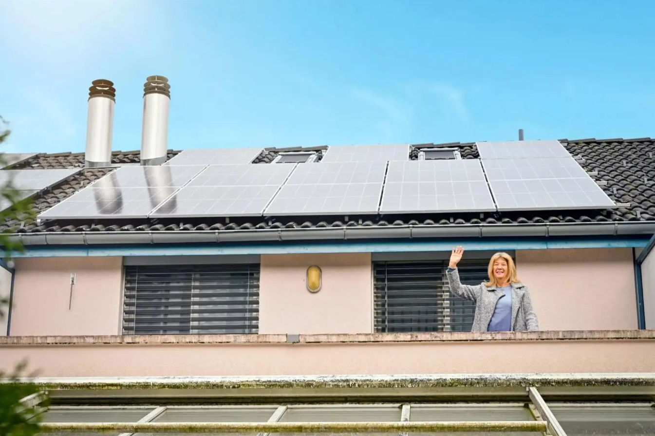 Frau auf Balkon mit Solaranlagen auf dem Dach, begrüßt die Kamera, blauer Himmel im Hintergrund.
