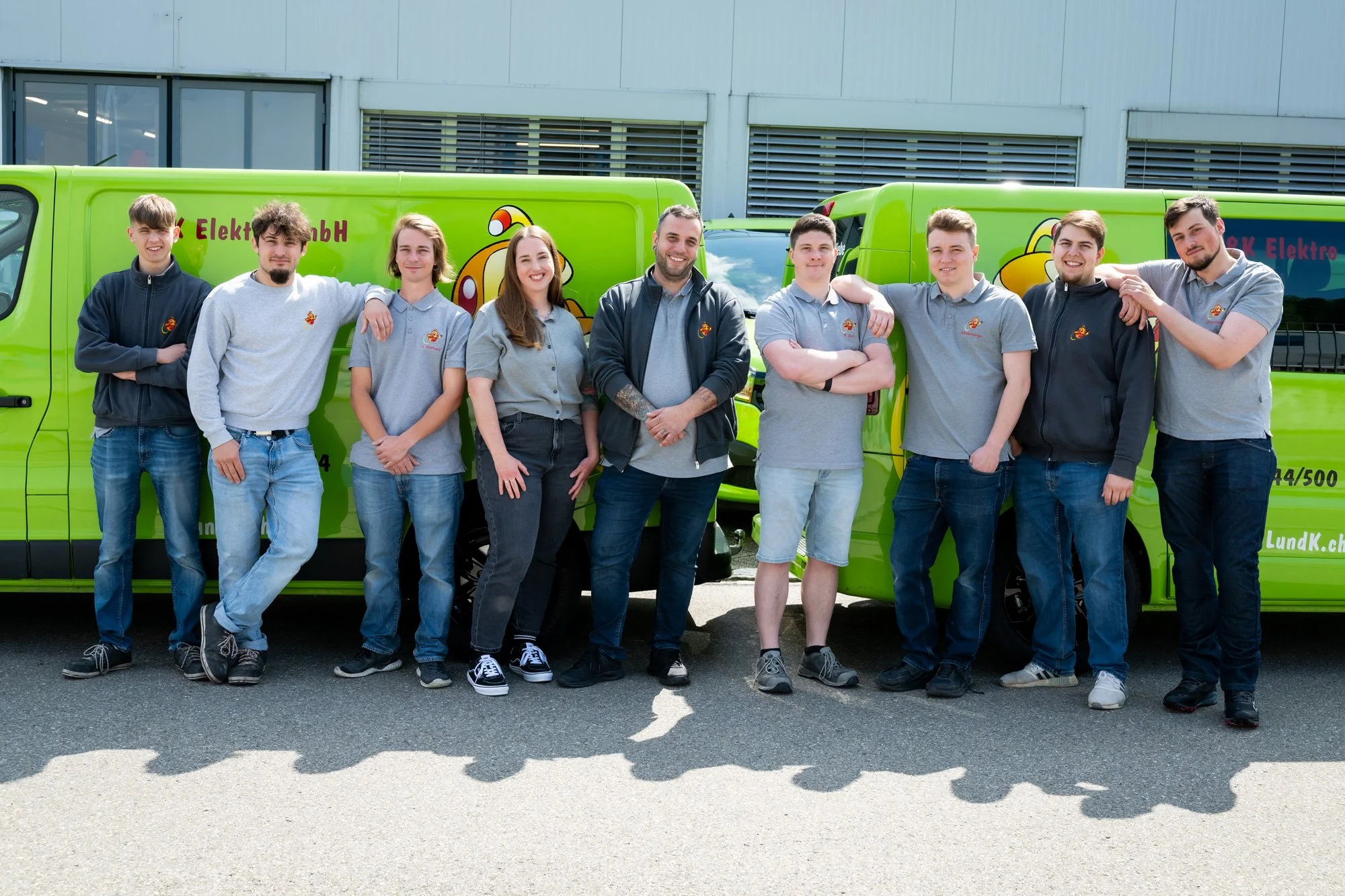 Gruppe von neun Menschen steht vor einem grünen Fahrzeug mit Laptop, im Hintergrund ein Gebäude