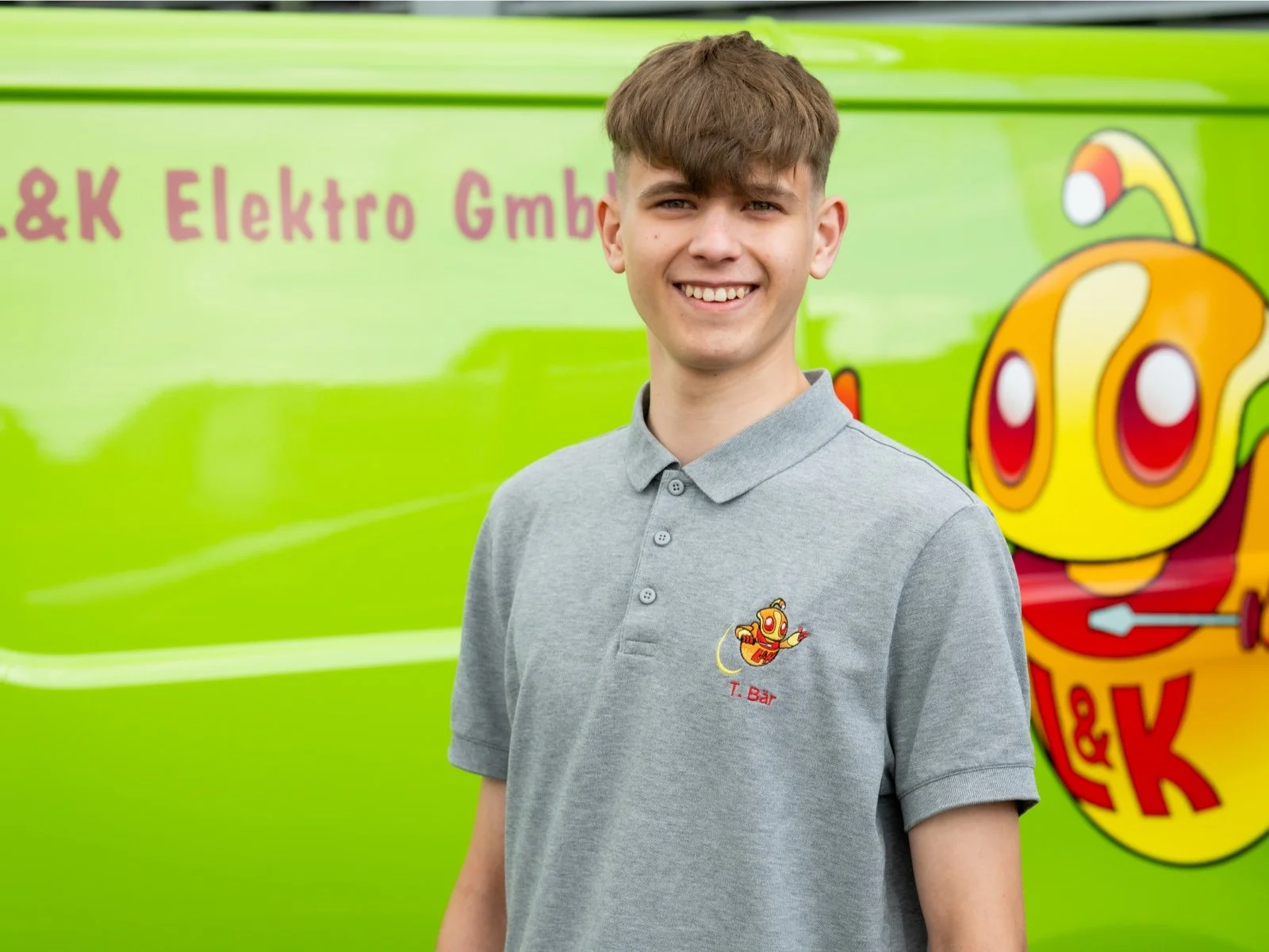 Junge trägt ein graues T-Shirt mit Logo einer Firma, steht vor einer grünen Firmenwagen mit buntem Logo und Beschriftung.