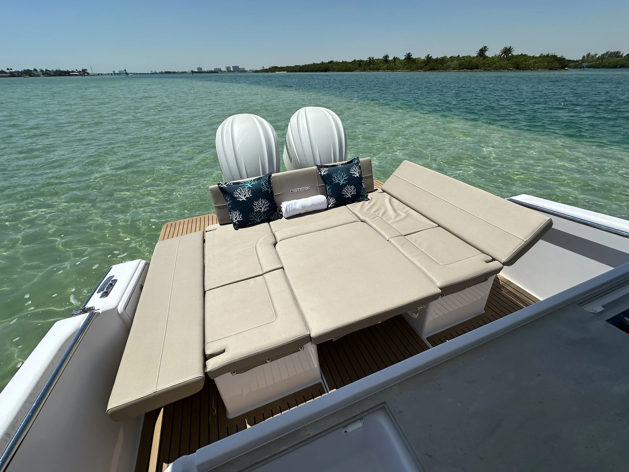 A yacht with a beige cushioned sunbed, two black pillows with white coral patterns, a white rolled towel, and two white outboard engines, floating on clear blue water near a lush green island with palm trees in the background.