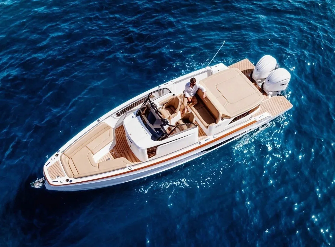 A white yacht with beige seating and twin engines floating on deep blue water, with a man in a white shirt sitting on the deck.