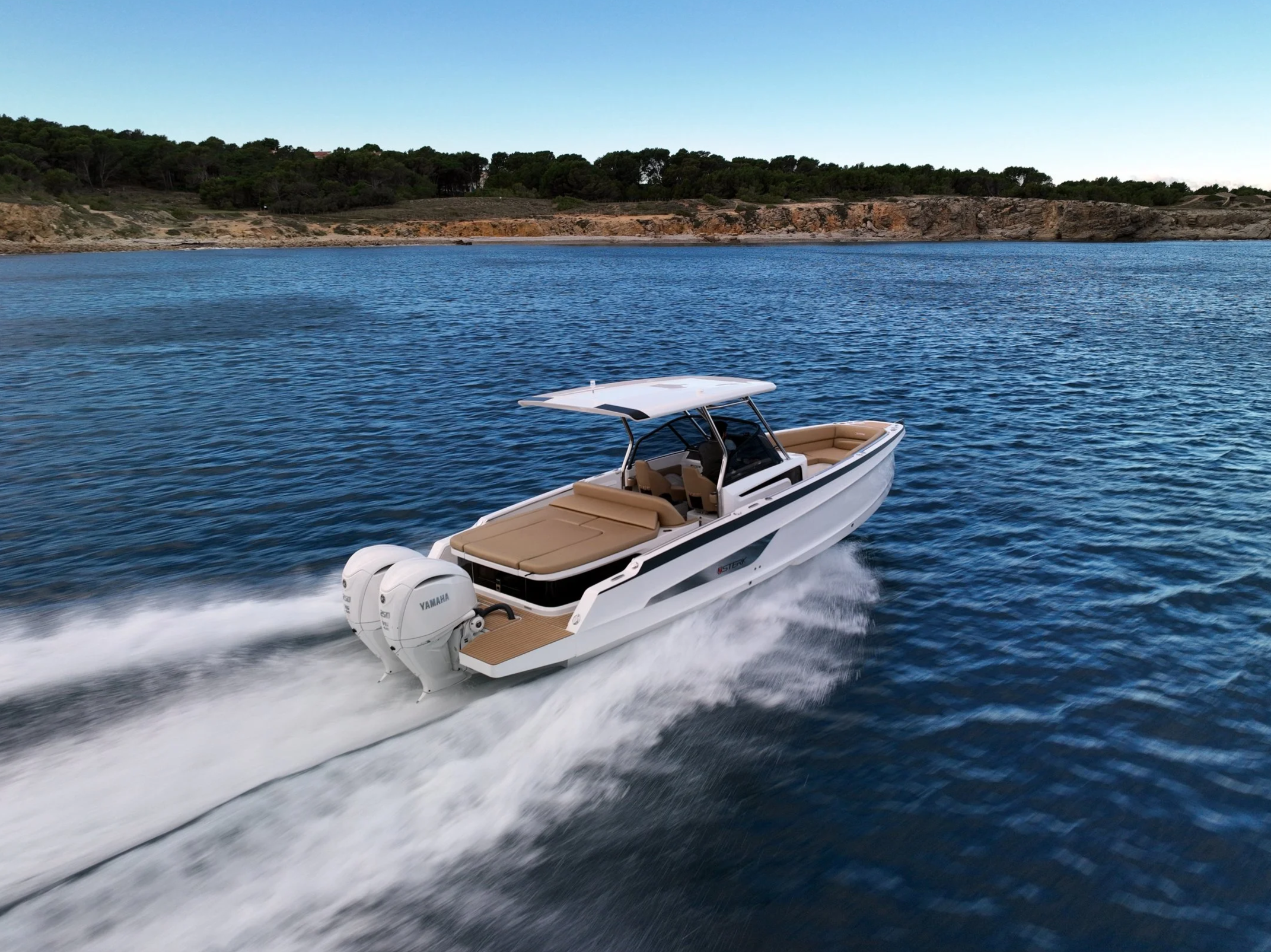 A white motorboat with beige interior cruising on a blue lake, leaving a wake behind, with a rocky and wooded shoreline under a clear sky in the background.
