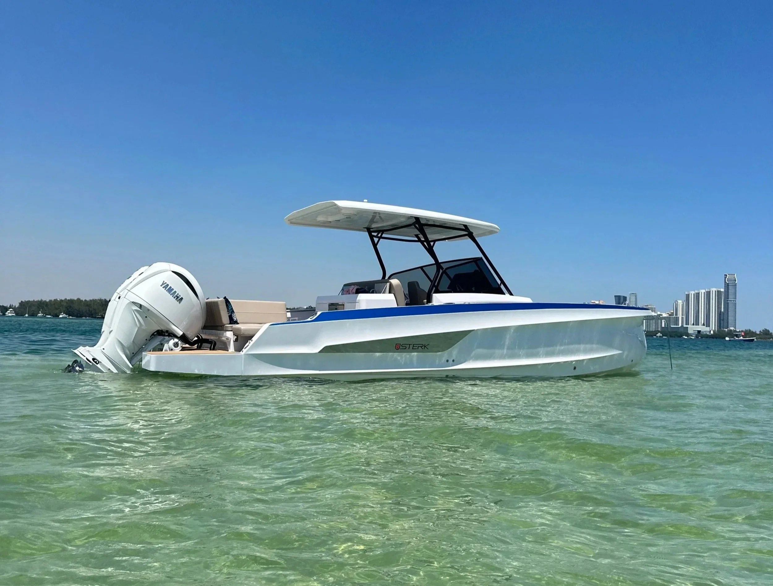 A small motorboat with a Yamaha outboard engine floating on clear, shallow water near a city skyline with tall modern buildings under a bright blue sky.