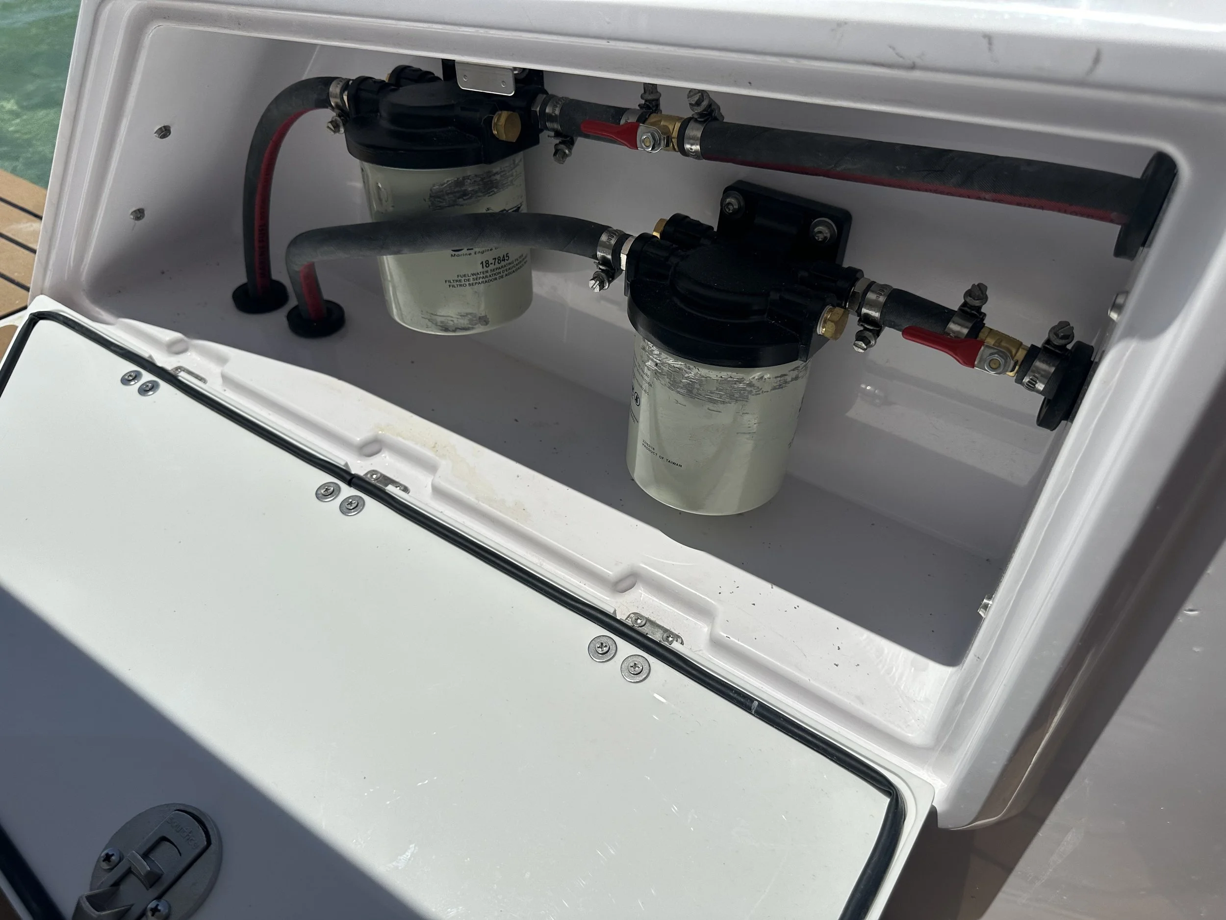 Inside a boat's compartment showing two fuel filters connected with hoses, and a closed white access panel with screws, with water visible in the background.