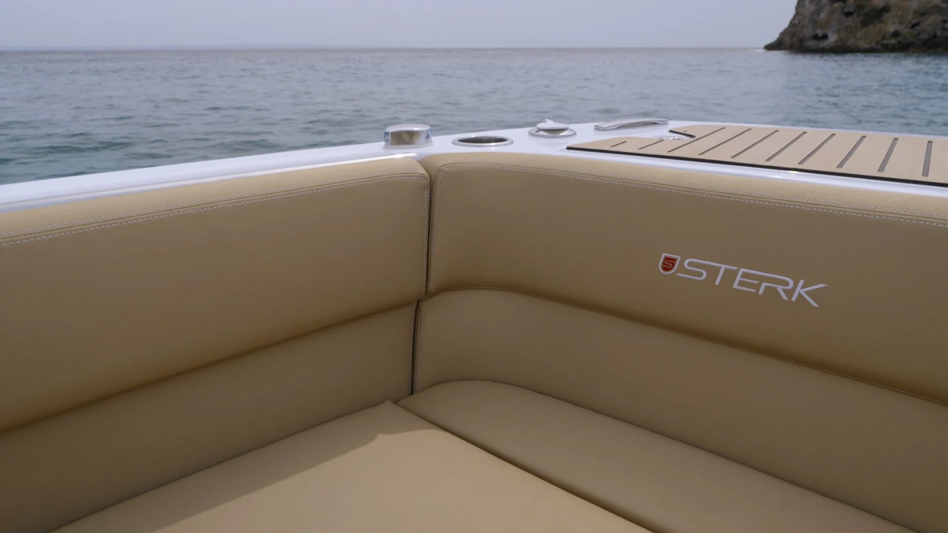 Close-up of beige upholstery on a boat with the logo 'STERK' and a background of the ocean and a rocky coastline.