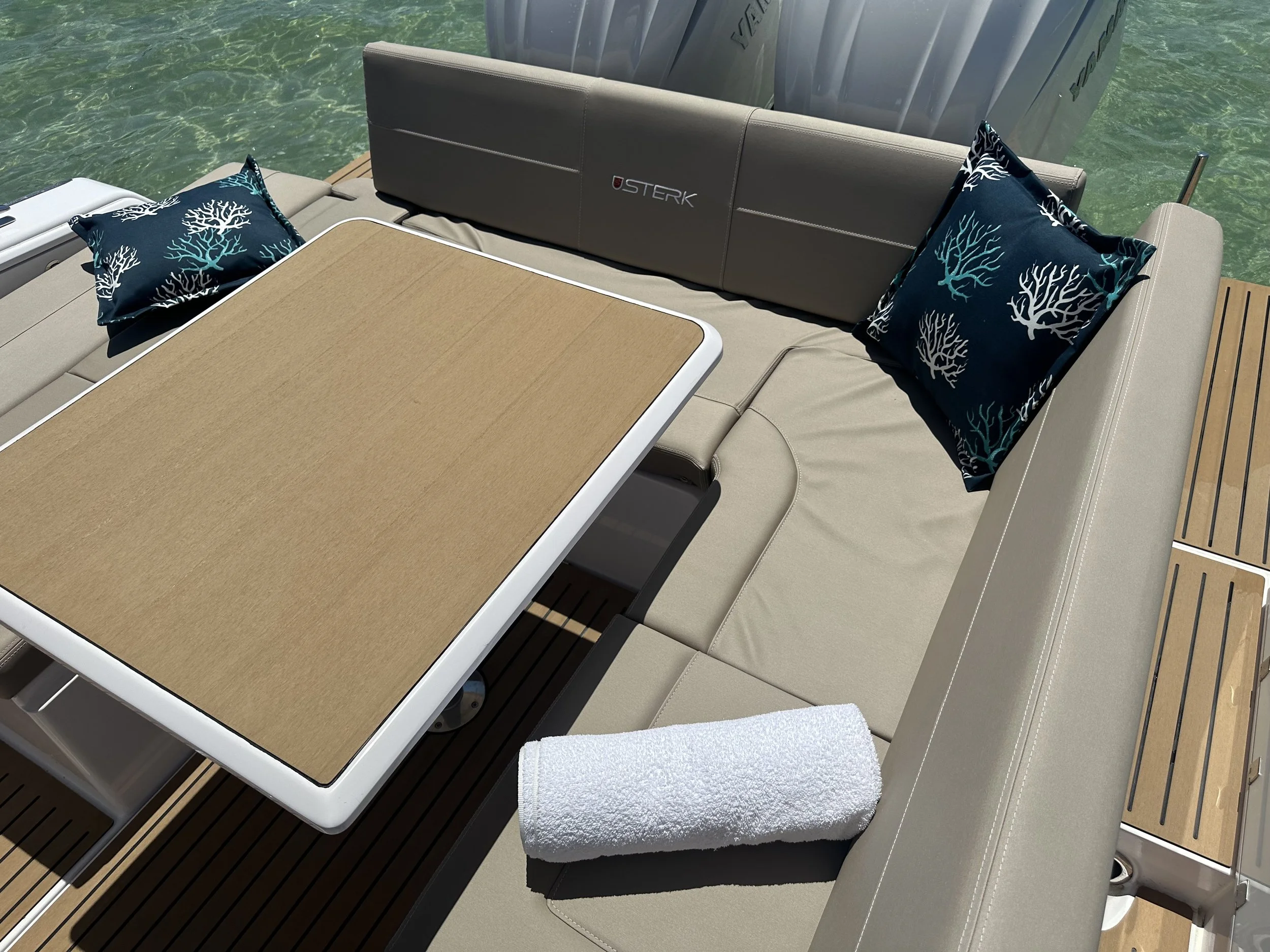 A boat seating area with a beige cushioned bench, two dark blue pillows with white coral patterns, a rolled white towel, a wooden table, and a water background.