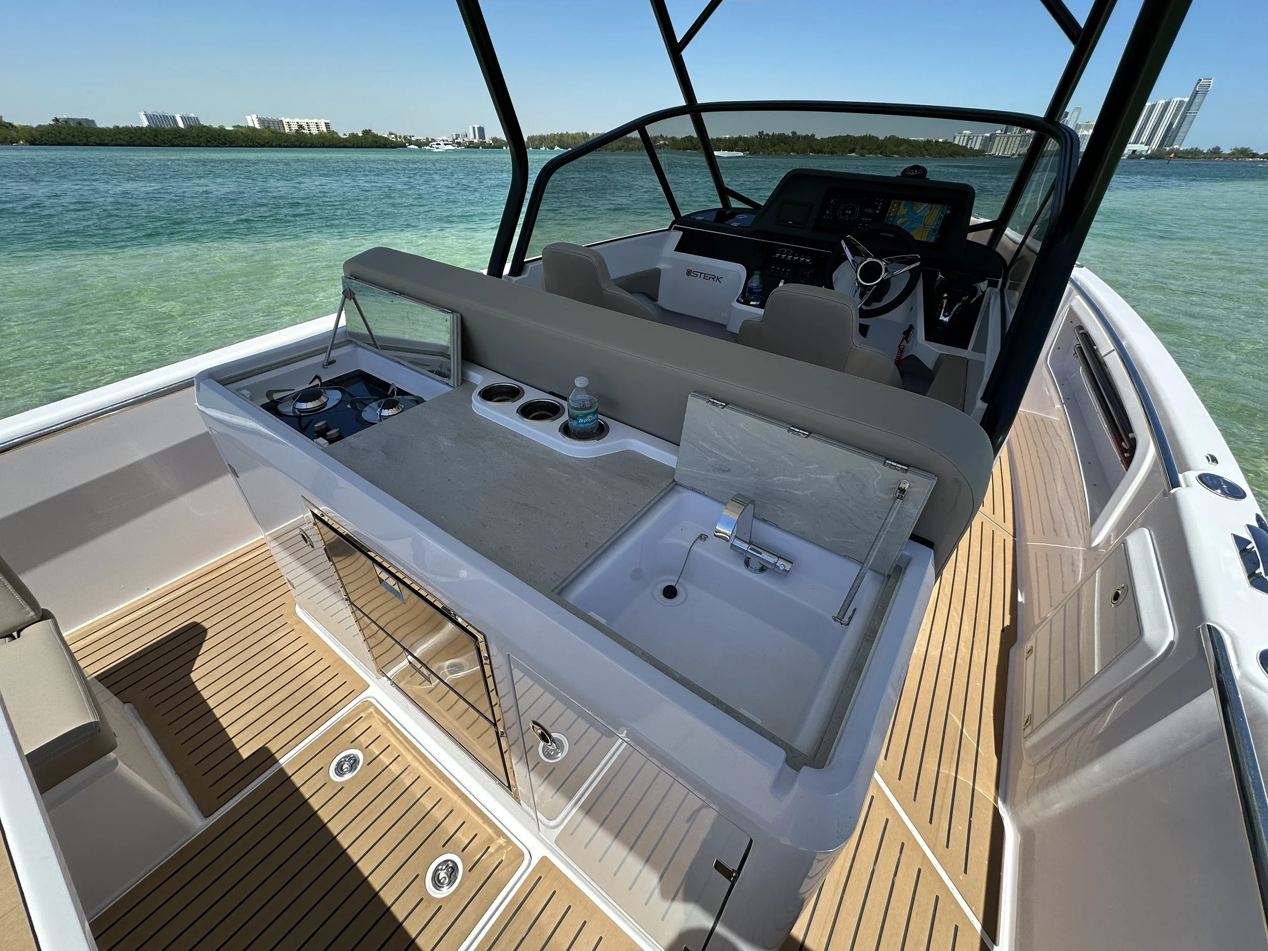 View of a luxury boat with a wood deck, a small sink, a stove, and a water bottle, on calm water with a cityscape in the background.