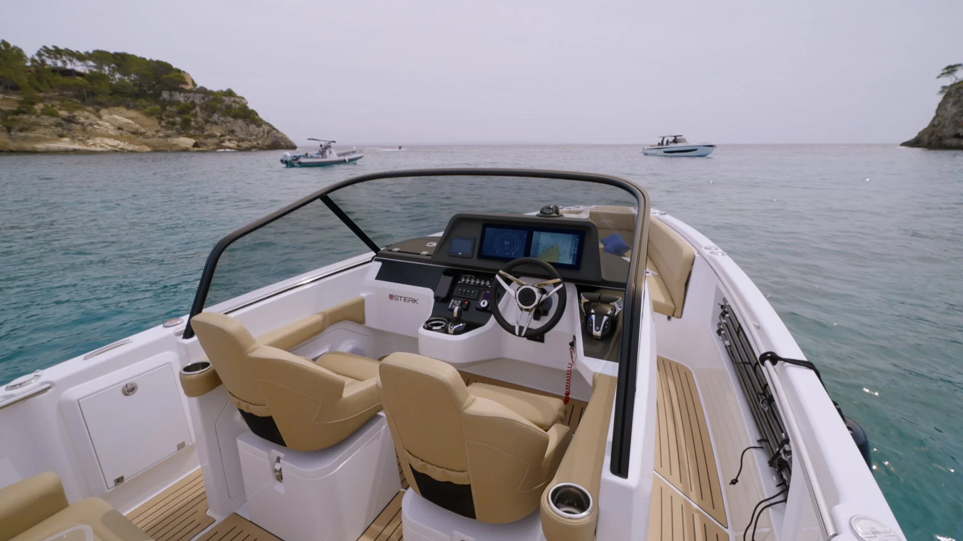 View from the cockpit of a luxury yacht, facing the helm with the steering wheel and navigation screens, overlooking calm water with other boats and rocky hills in the distance.