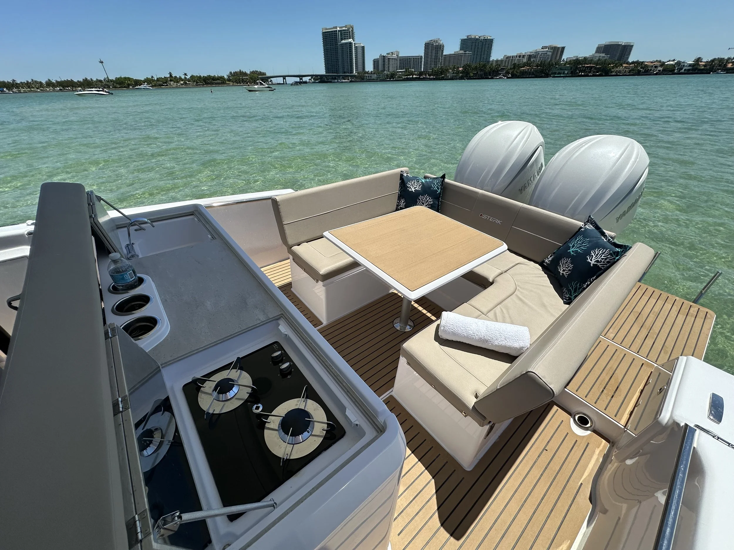 Seating area with cream-colored cushioned chairs and cushions, a wooden table, and a built-in kitchenette with stovetop on a boat deck. Twin outboard motors and a cityscape with high-rise buildings are visible in the background.