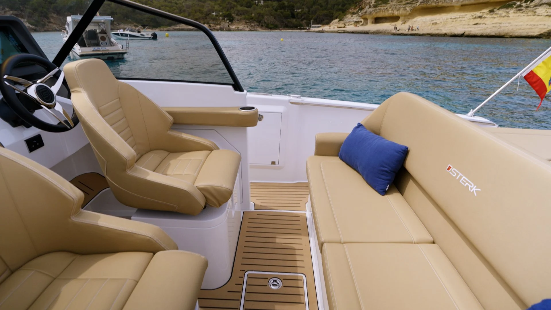 Interior view of a boat with beige leather seats, a blue pillow on the right seat, and a view of the water and boats outside. Sterk 31RC cockpit