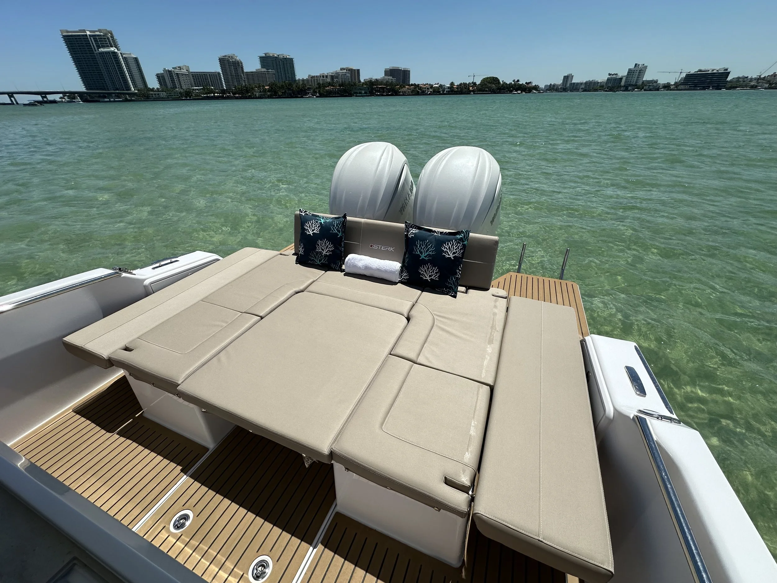 View of a boat with beige lounge cushions, black pillows with coral patterns, and two outboard motors on a body of water with a city skyline in the background.