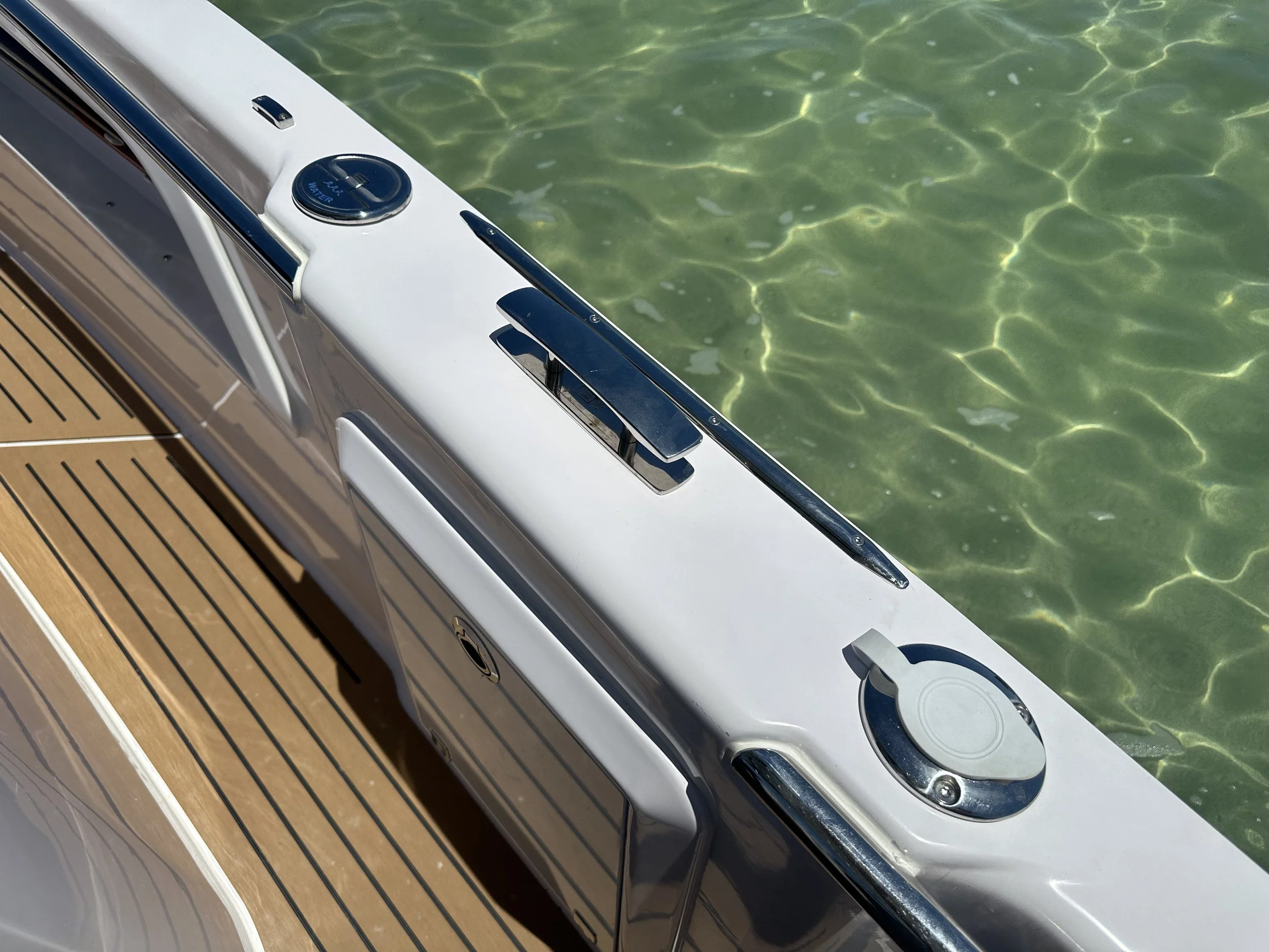 Close-up of a boat's deck with a white console, stainless steel fittings, and a wooden deck, floating on clear green water.