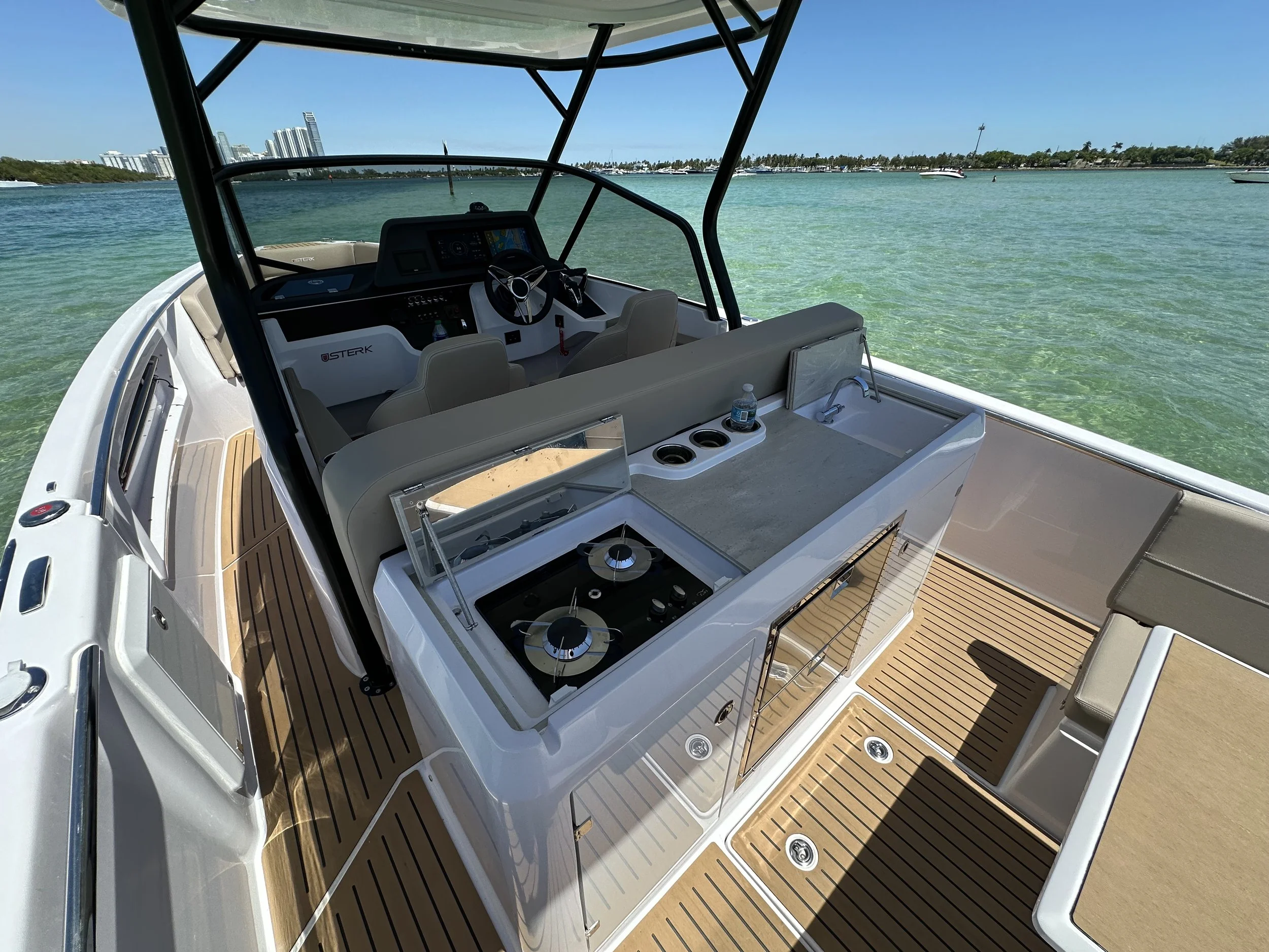 View of a modern boat with a wooden deck on the water. The boat has a steering wheel, navigation system, and a small kitchen area with a stove and sink. The background shows a city skyline and a clear blue sky.