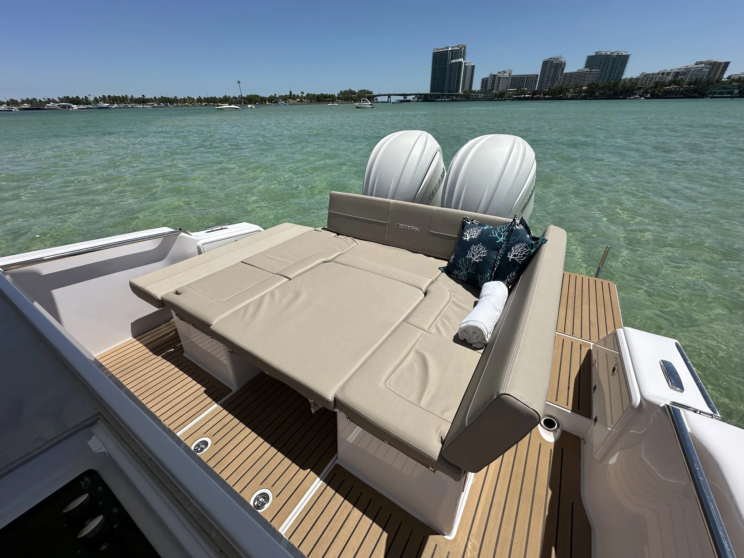 A boat deck with a beige cushioned sunbed, two decorative pillows with ocean motifs, a rolled white towel, and two white outboard motors in the water with a city skyline and clear blue sky in the background.