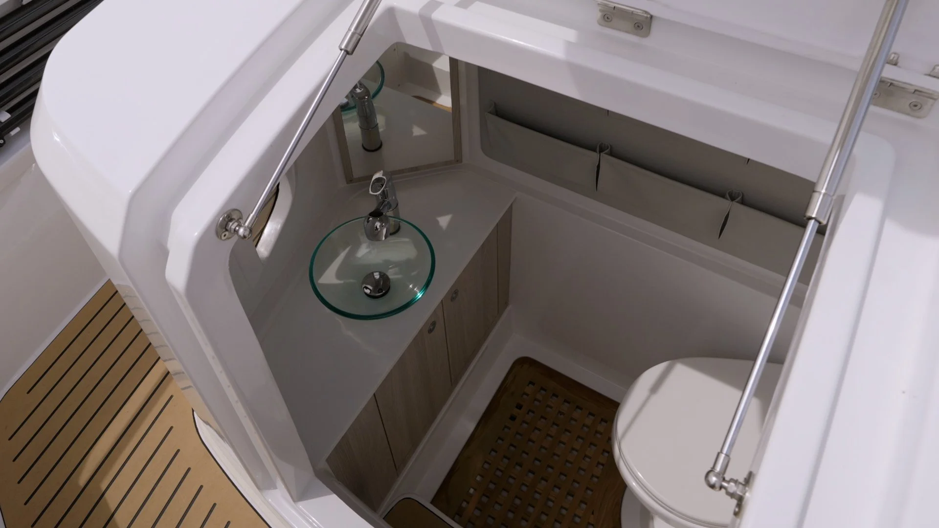 Interior of a yacht bathroom featuring a small glass sink with a chrome faucet, wooden cabinetry, a mirror, and a small white toilet, with a rubber mat on the floor. Sterk 31 heads