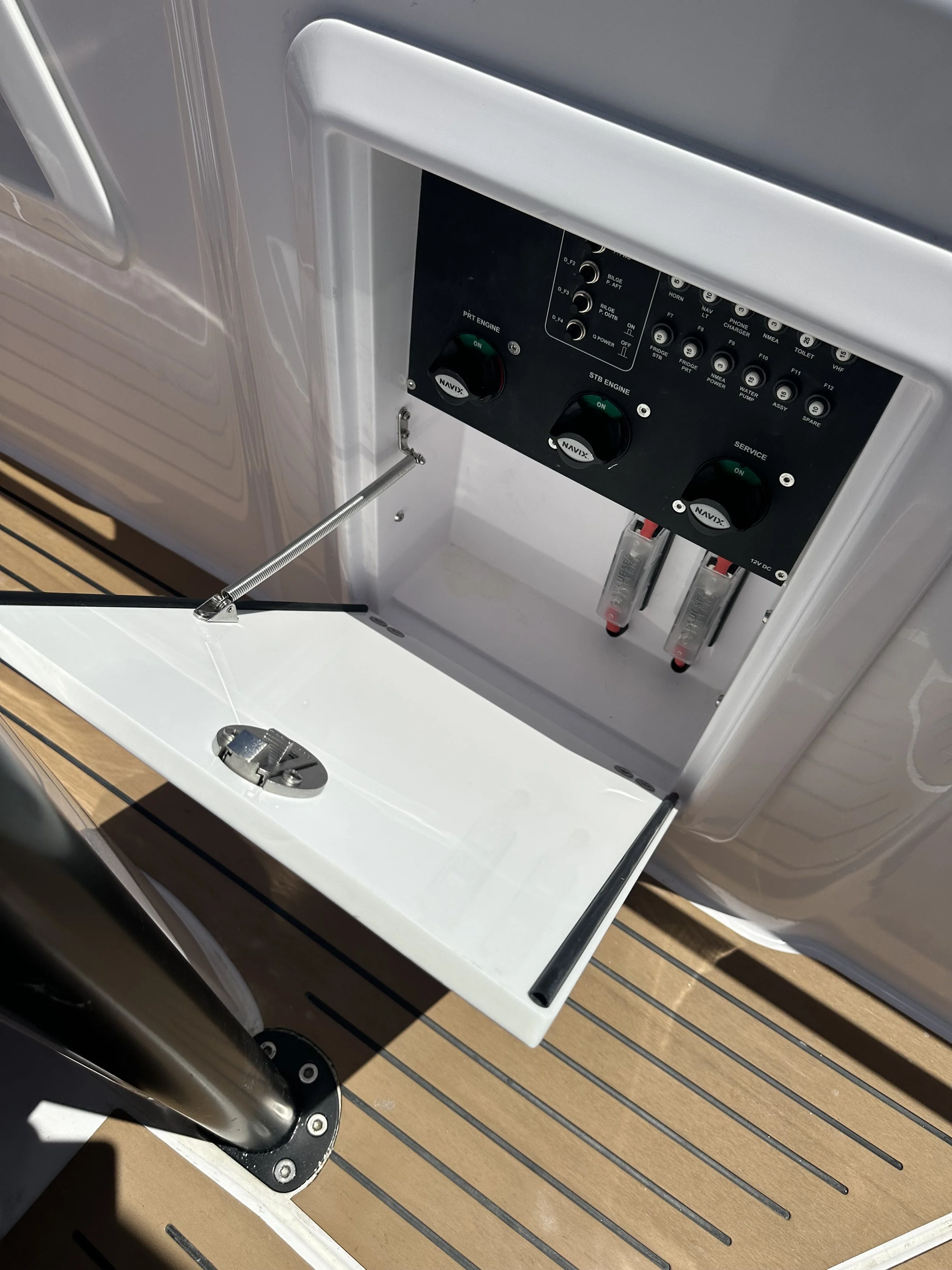 Electrical control panel with switches and circuit breakers on a boat, with a white service hatch open below the panel.