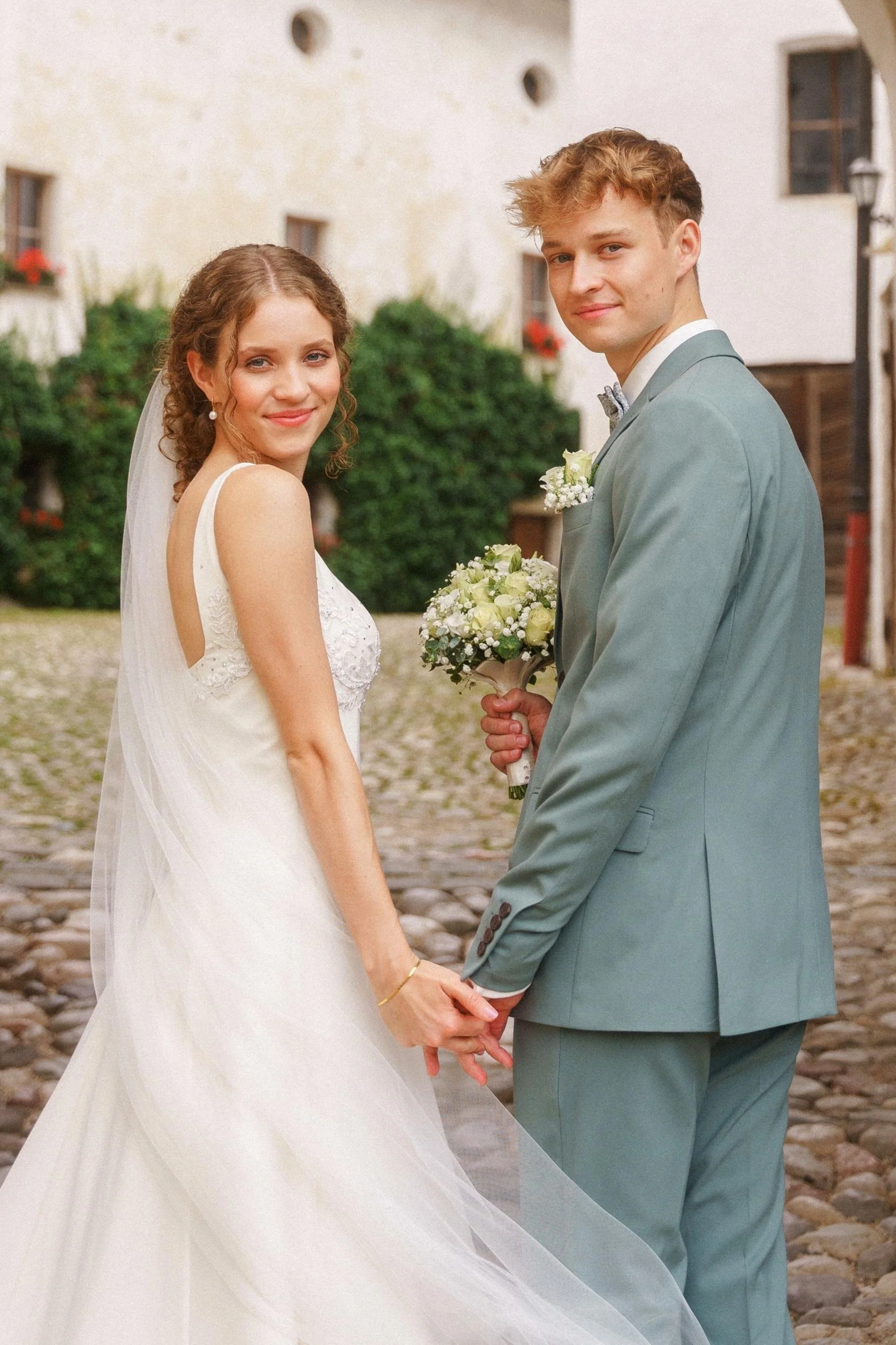 Ein frisch verheiratetes Paar, der Mann trägt einen hellblauen Anzug und die Dame ein weißes Kleid mit Schleier, der Mann hält einen Brautstrauß mit weißen Blumen