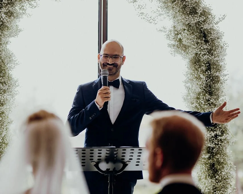 Ein Mann in einem schwarzen Anzug und mit Fliege hält eine Hand aus und spricht bei einer Hochzeitsfeier, während er ein Mikrofon hält.
