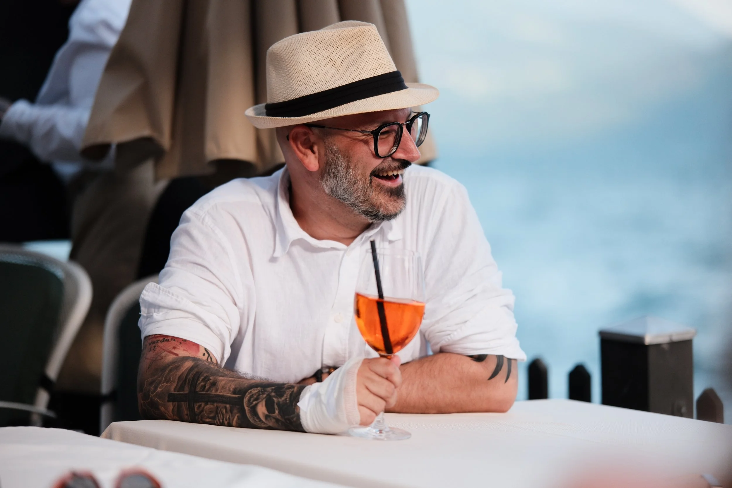 Lächelnder Mann mit Brille, Sonnenhut und Tattoo, der ein orangefarbenes Getränk mit Strohhalm in der Hand hält, bei einem Treffen am Tisch im Freien mit Wasser im Hintergrund.