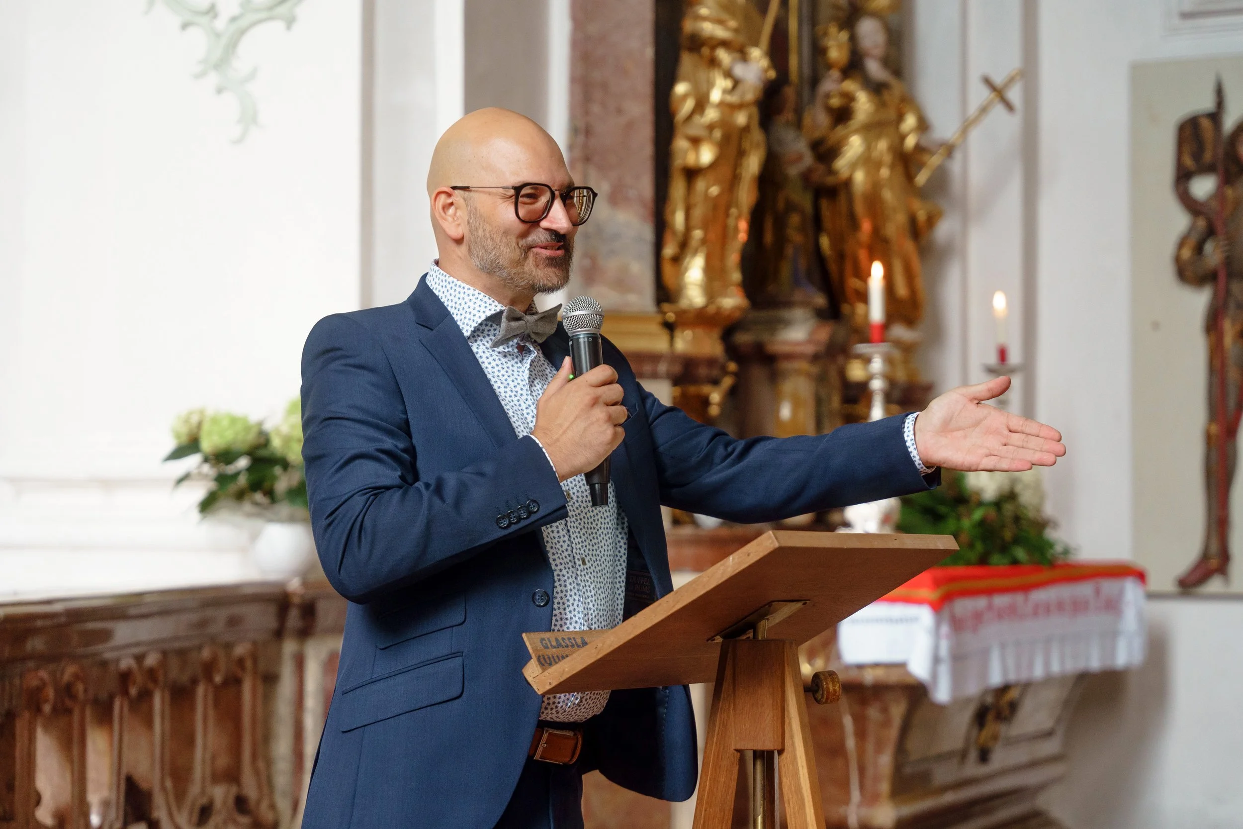 Ein Mann in formeller Kleidung, der an einer Hochzeitsmesse spricht, während er eine Hand ausstreckt, um seine Rede zu betonen, im Hintergrund religiöse Kunst und Kerzen in einer Kirche.