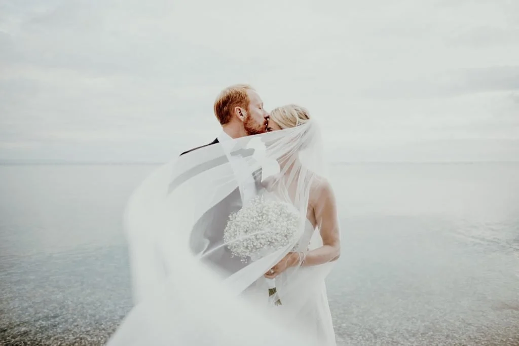 Ein Brautpaar, das sich an einem ruhigen Strand küsst, bei bewölkten Himmel, mit dem Meereshintergrund.