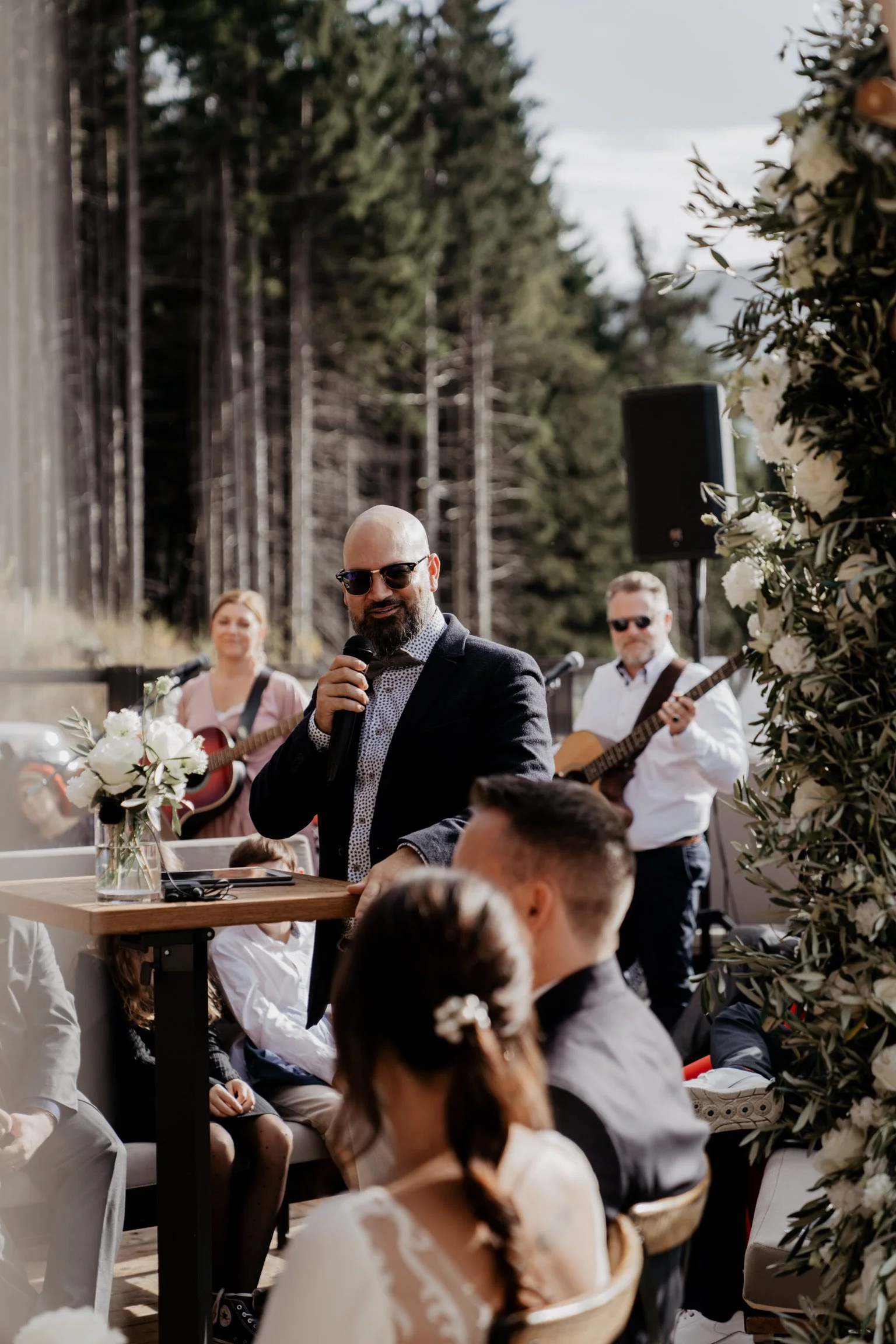 Mann mit dunklem Anzug und Sonnenbrille spricht in Mikrofon bei Hochzeitsfeier im Freien, im Hintergrund spielen zwei Musiker auf Gitarren, umgeben von weißen Blumen und einer Waldkulisse.