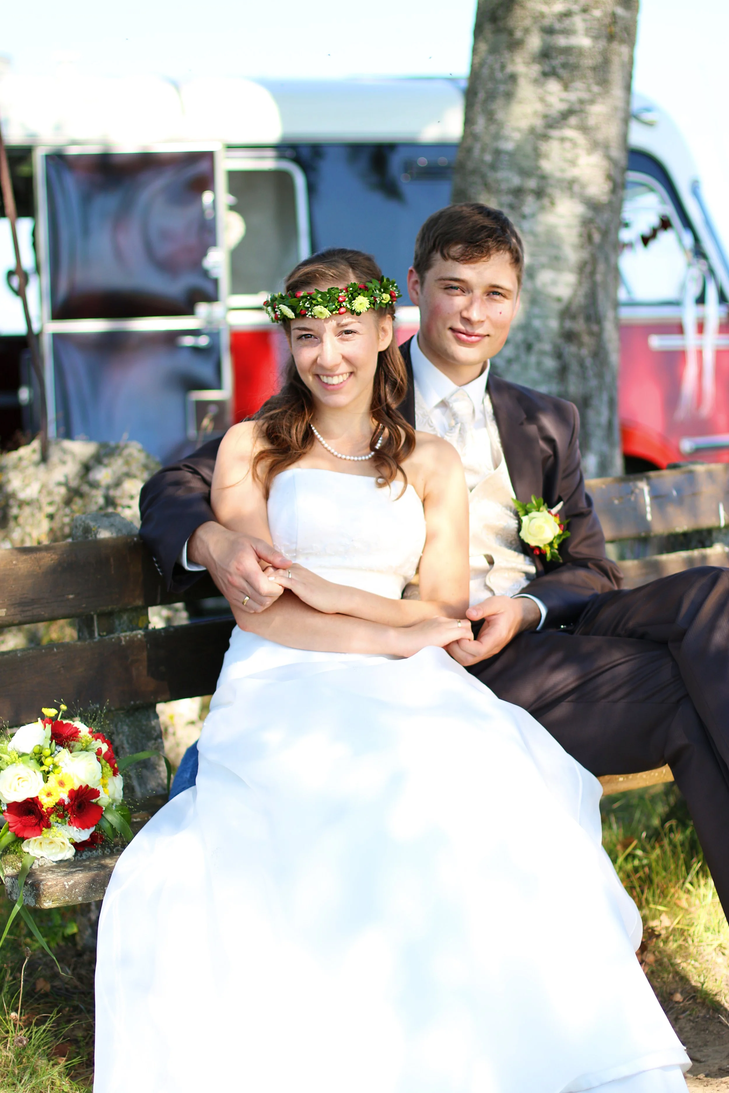Ein frisch verheiratetes Paar sitzend auf einer Parkbank, im Freien, mit Blumen und einem klassischen Fahrzeug im Hintergrund, bei Tageslicht.
