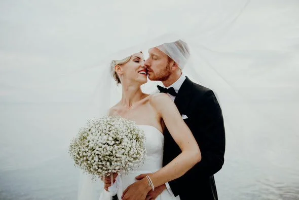 Frisch verheiratetes Paar, die Dame hält einen Hochzeitsstrauß mit weißen Blumen, trägt ein weißes Kleid und der Mann einen schwarzen Anzug, der Hintergrund ist Wasser und ein weißer Schleier umhüllt das Bild