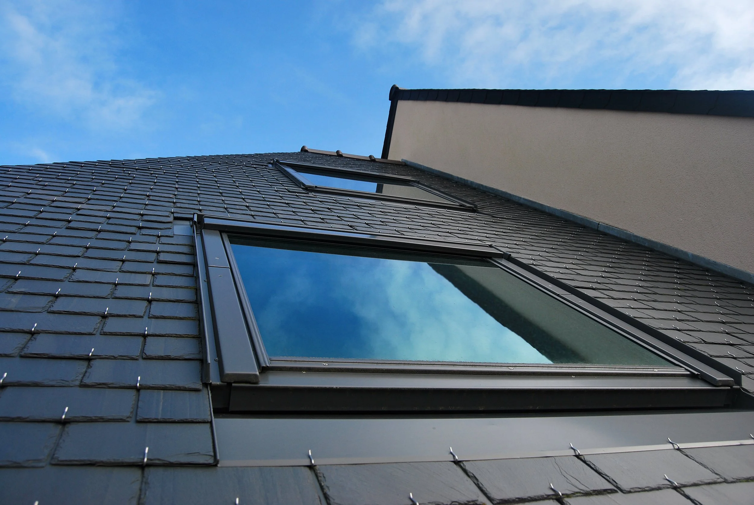 Low-angle view of a modern building with black slate roof tiles and a large glass skylight, against a blue sky with some clouds.
