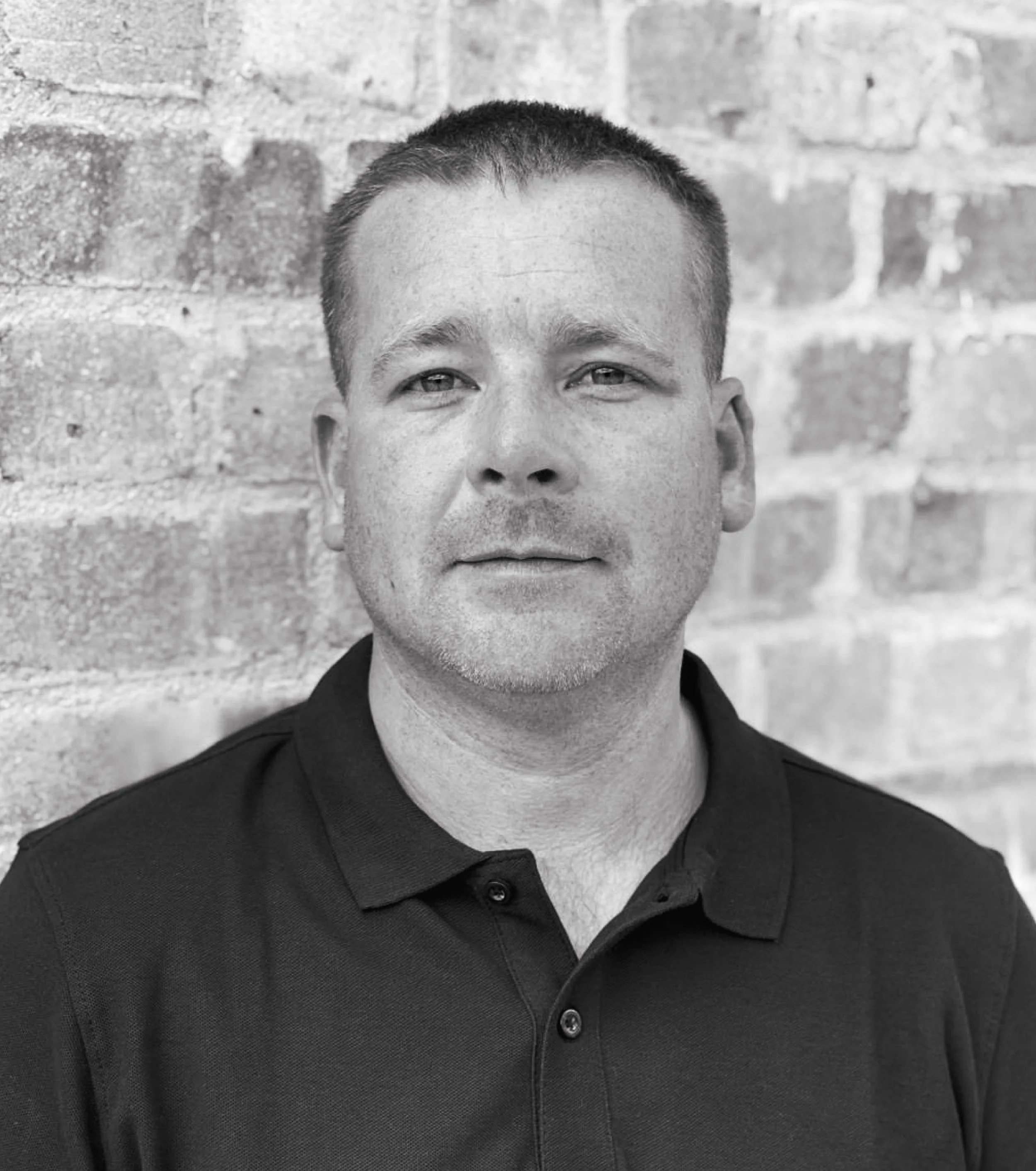 Black and white portrait of a man with short hair, wearing a collared shirt, standing in front of a brick wall.