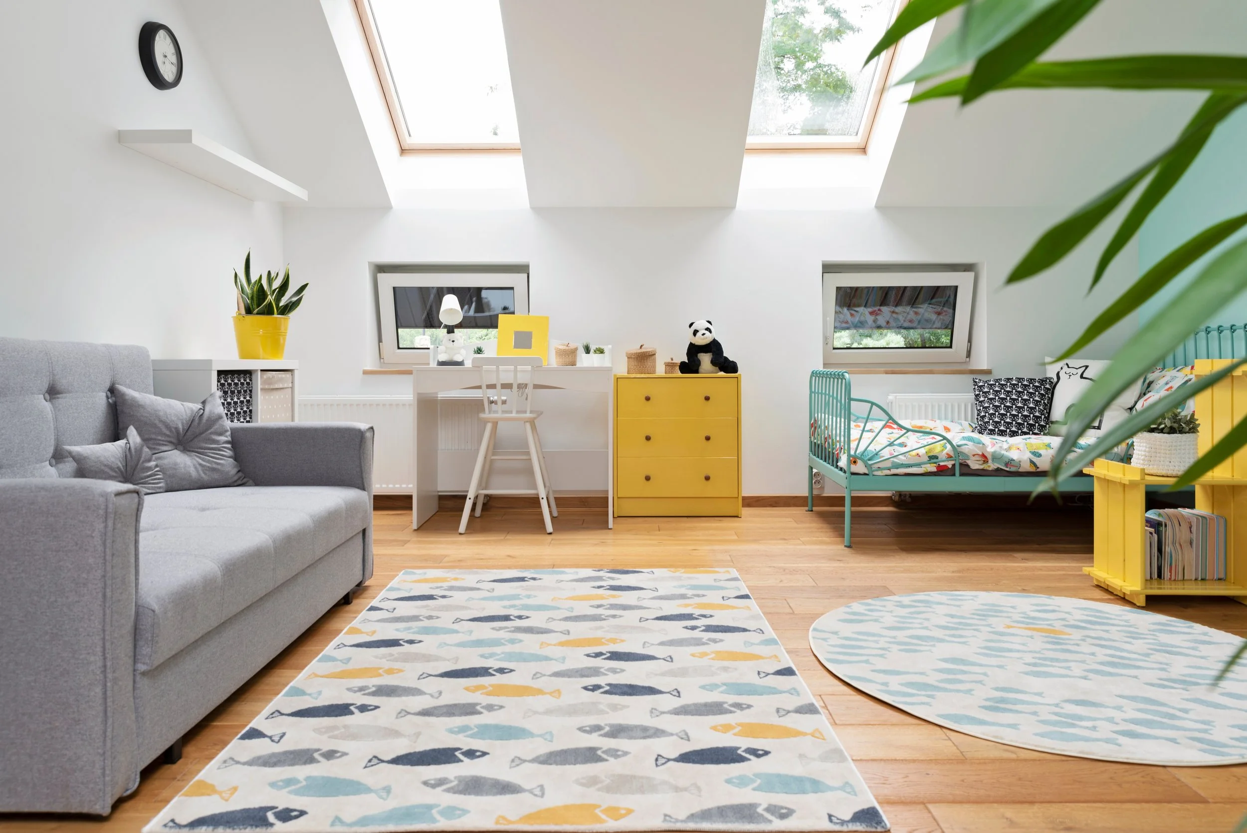 Bright attic room with sloped ceiling and skylights, white walls, wooden floor, containing a gray sofa with pillows, a yellow dresser with a panda plush, a white desk with a chair, a teal bed with patterned bedding and pillows, yellow storage unit wi