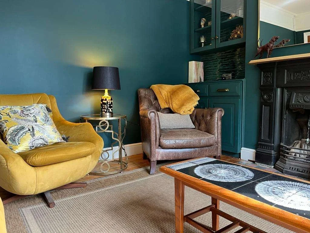 Living room with yellow sofa, brown armchair with throw blanket, green cabinet, black fireplace, and coffee table, decorated with various items and artwork.