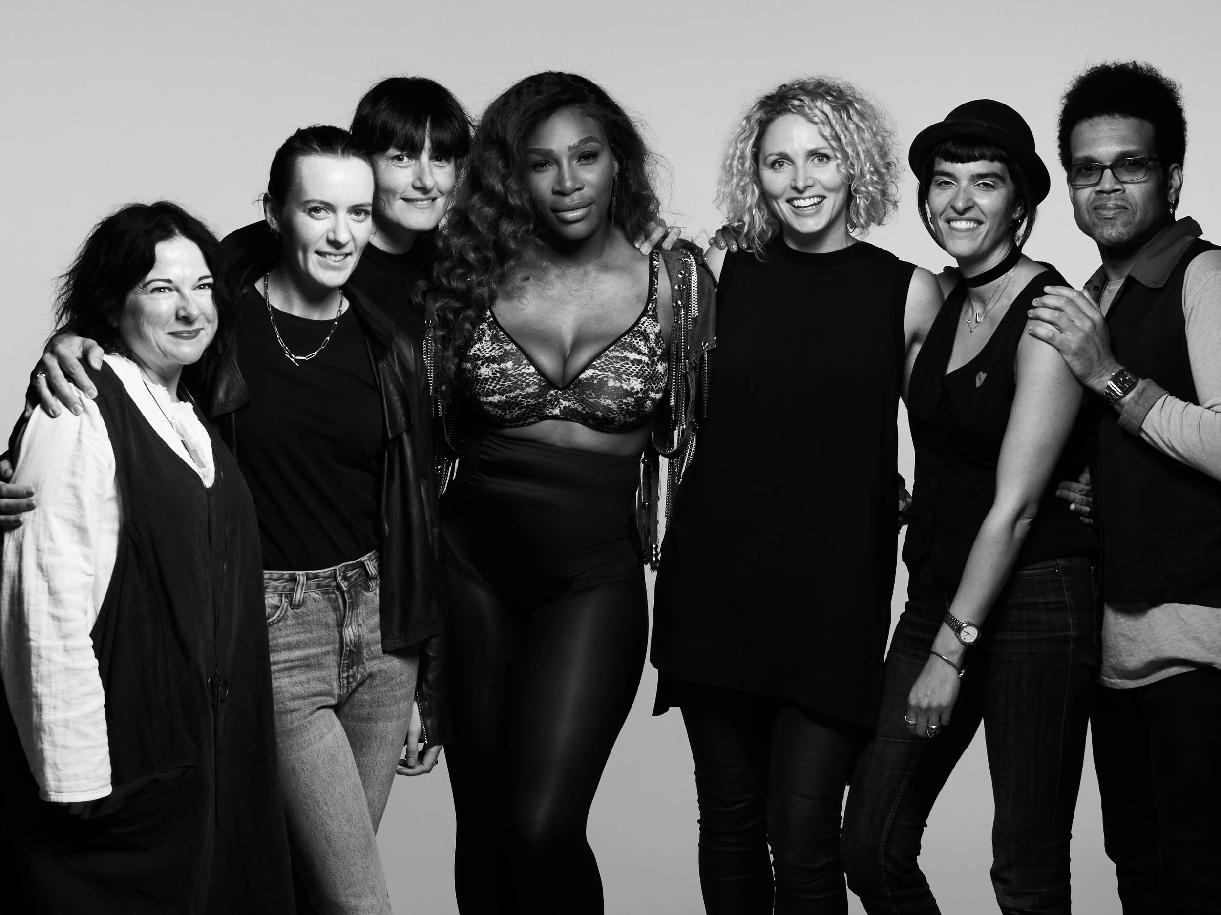 A black-and-white photo of eight diverse women and one man posing together. They are standing close with their arms around each other, smiling at the camera.