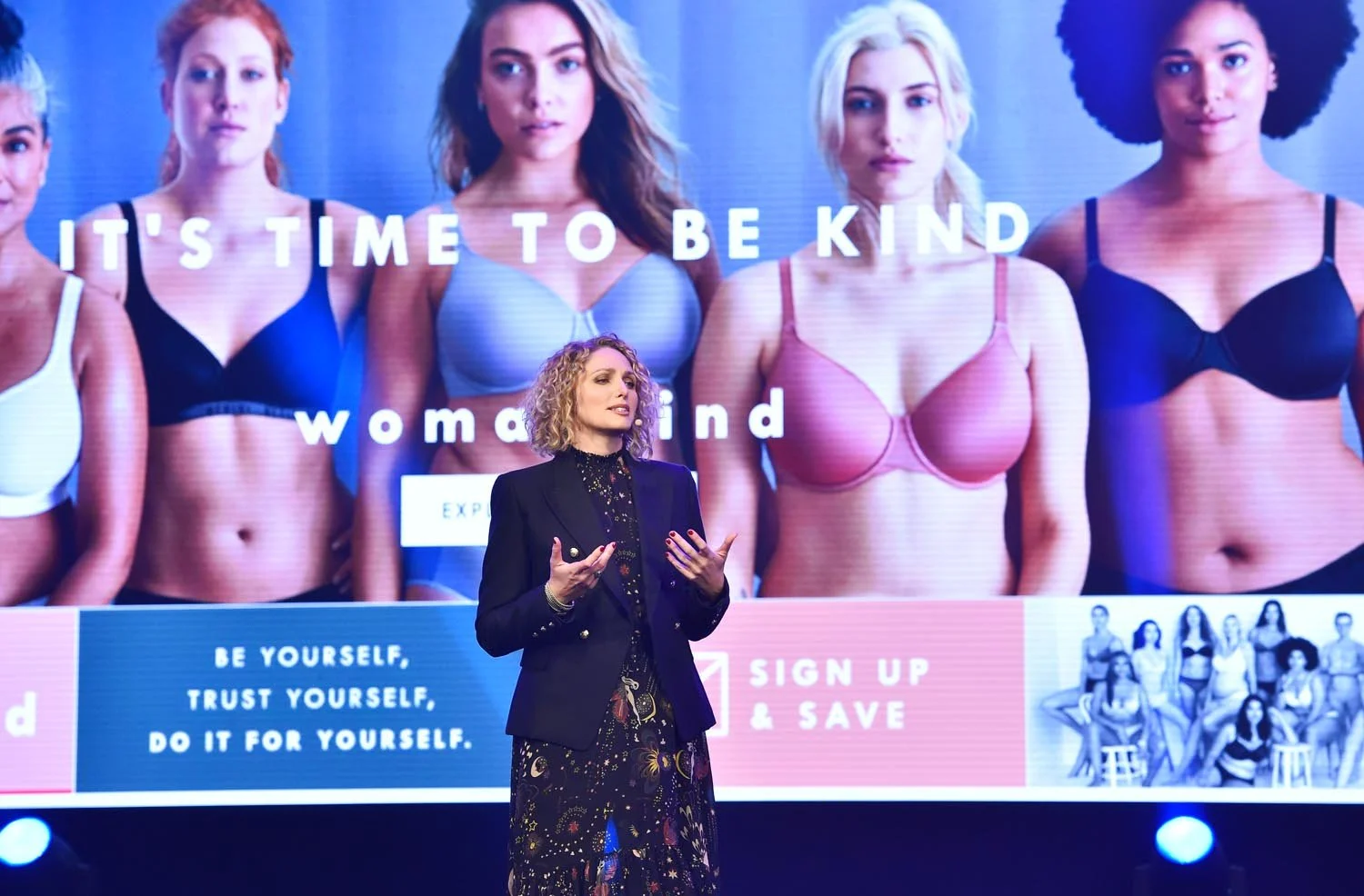 A woman with curly blonde hair giving a presentation in front of a large screen displaying diverse women in bras with the message 'It's time to be kind to women and'. The screen also shows options for signing up and saving, with a group photo of women in lingerie at the bottom right.