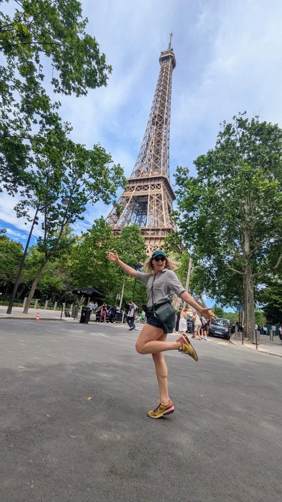 A woman joyfully posing in front of the Eiffel Tower in Paris, France on a clear day with blue skies and green trees.