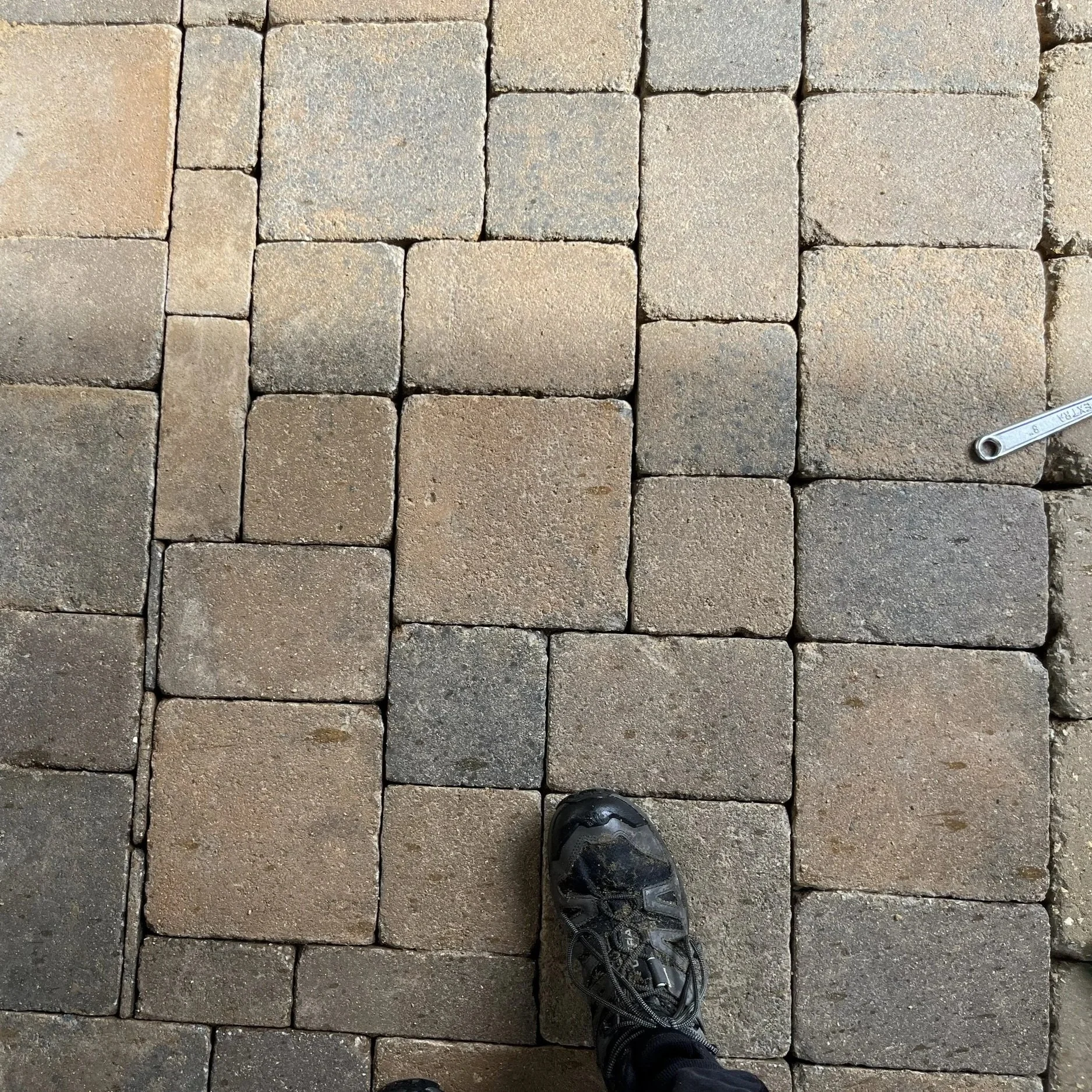 A person's foot wearing a black hiking shoe on a paved stone walkway with tan and gray bricks. A metal wrench is on the right side of the bricks.