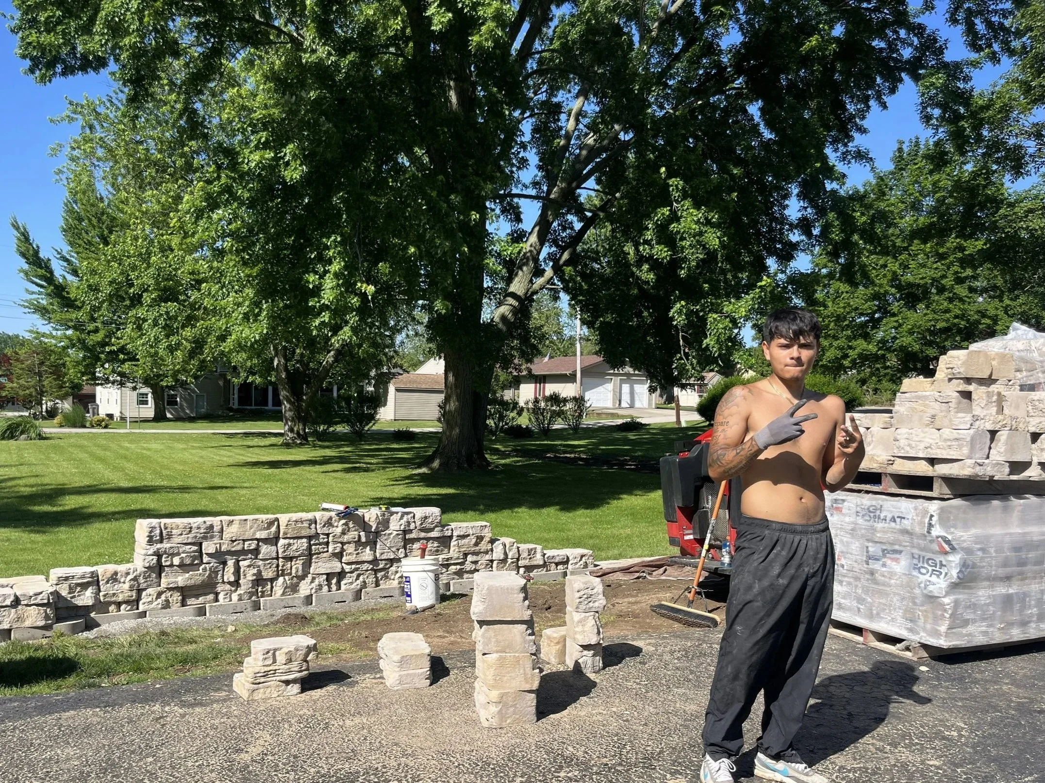 A shirtless man standing on a paved area in front of a brick wall, making a peace sign with his right hand, with construction materials and tools around him in a residential neighborhood with green trees and houses in the background.