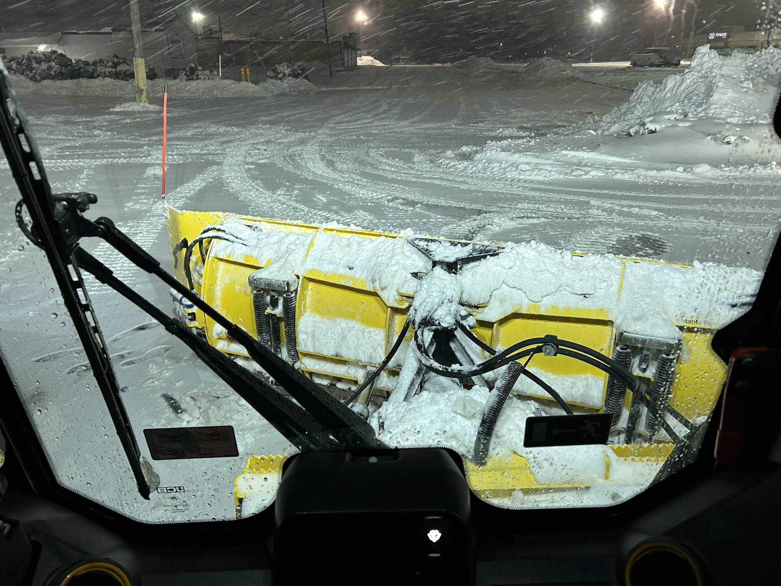 View from inside a snowplow cab showing the snowplow blade clearing snow from a street during a snowstorm at night.
