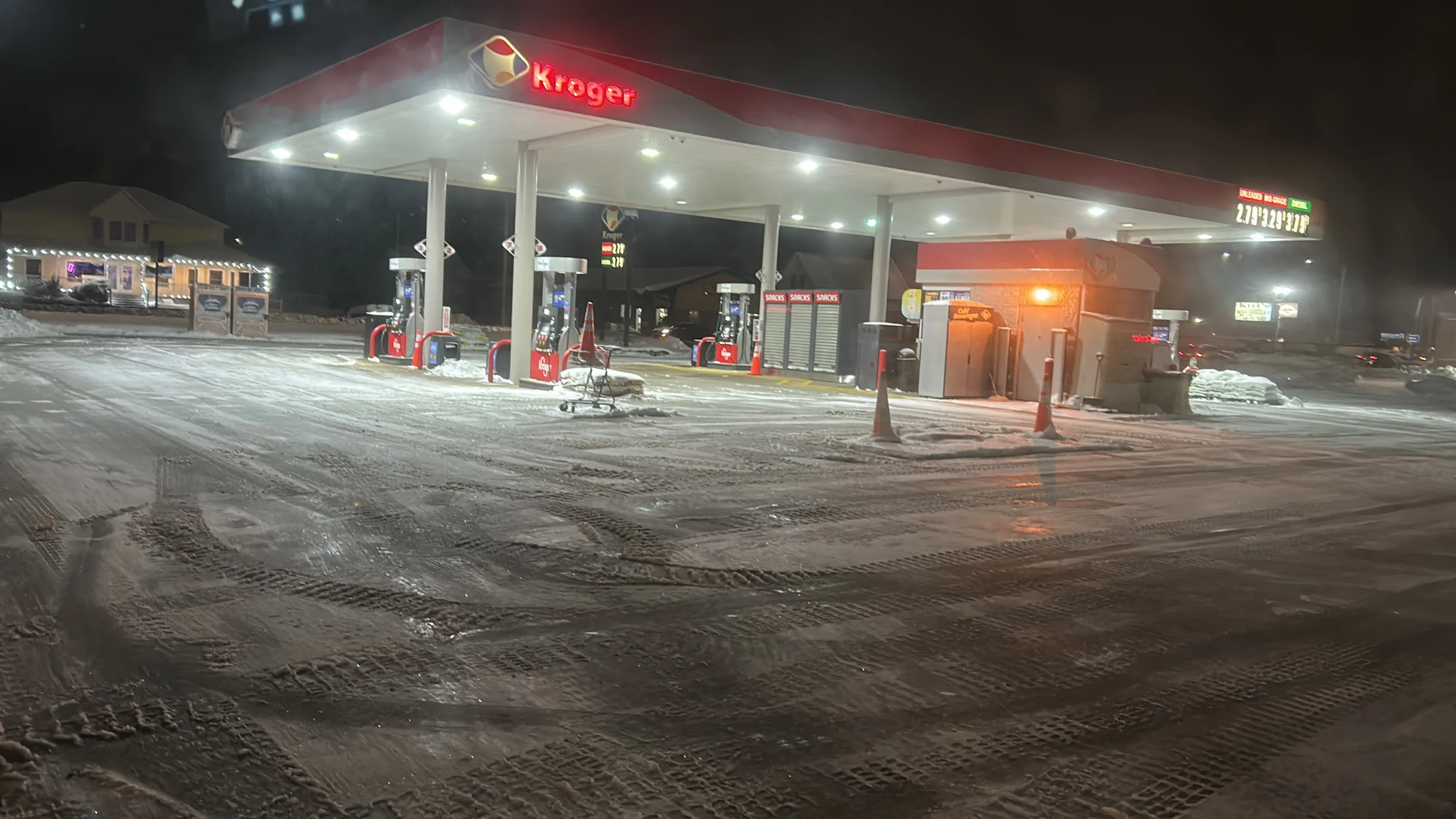 A snow-covered Kroger gas station at night with illuminated fuel pumps, a shopping cart in the snow, and a small building to the right. The ground has tire marks and snow, and there are trailers and buildings in the background.