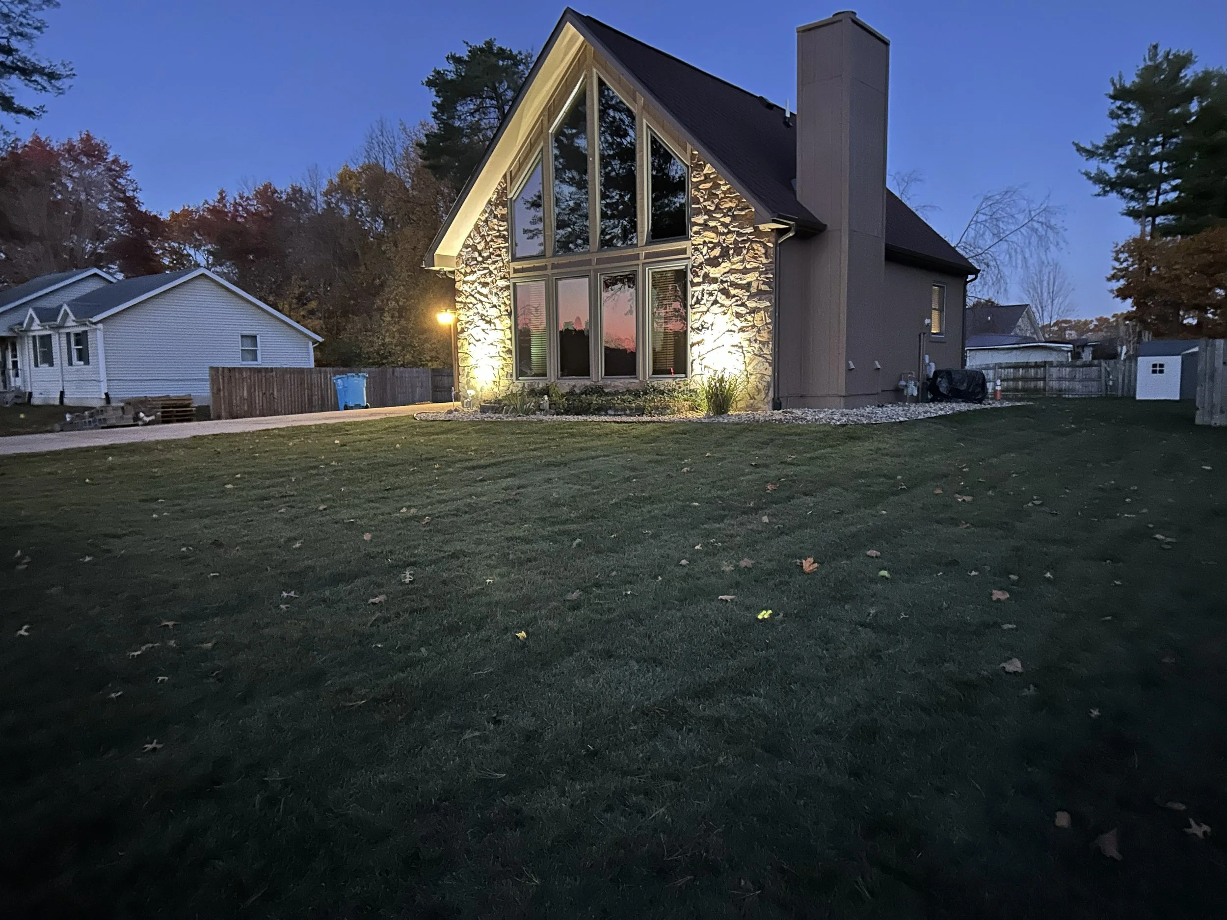 A modern house with large front windows, stone accent wall, and illuminated exterior lights, situated on a lawn during dusk with surrounding trees and neighboring houses.