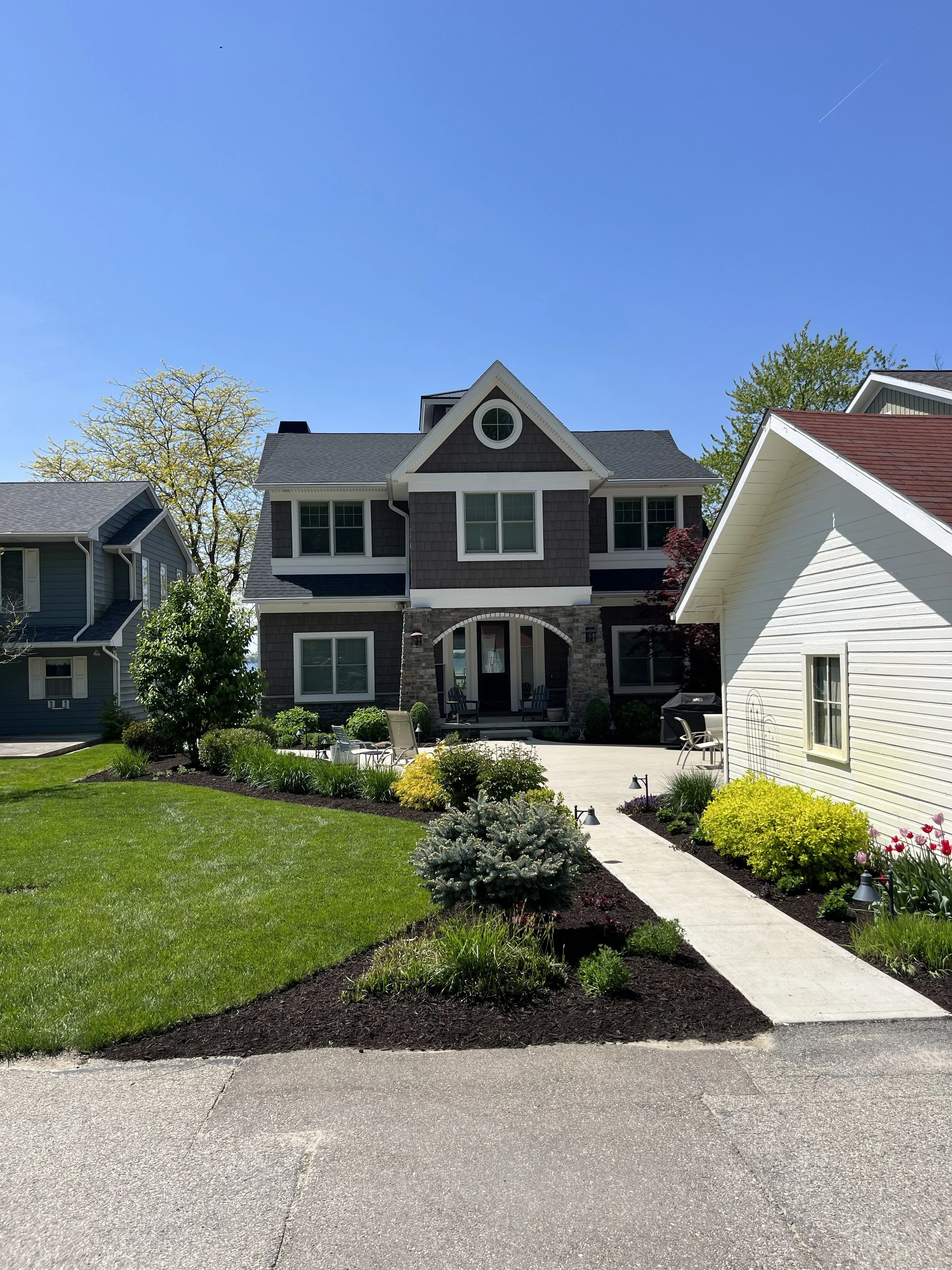 Front yard of a large house with well-manicured lawn, flower beds, and patio furniture, under a clear blue sky.