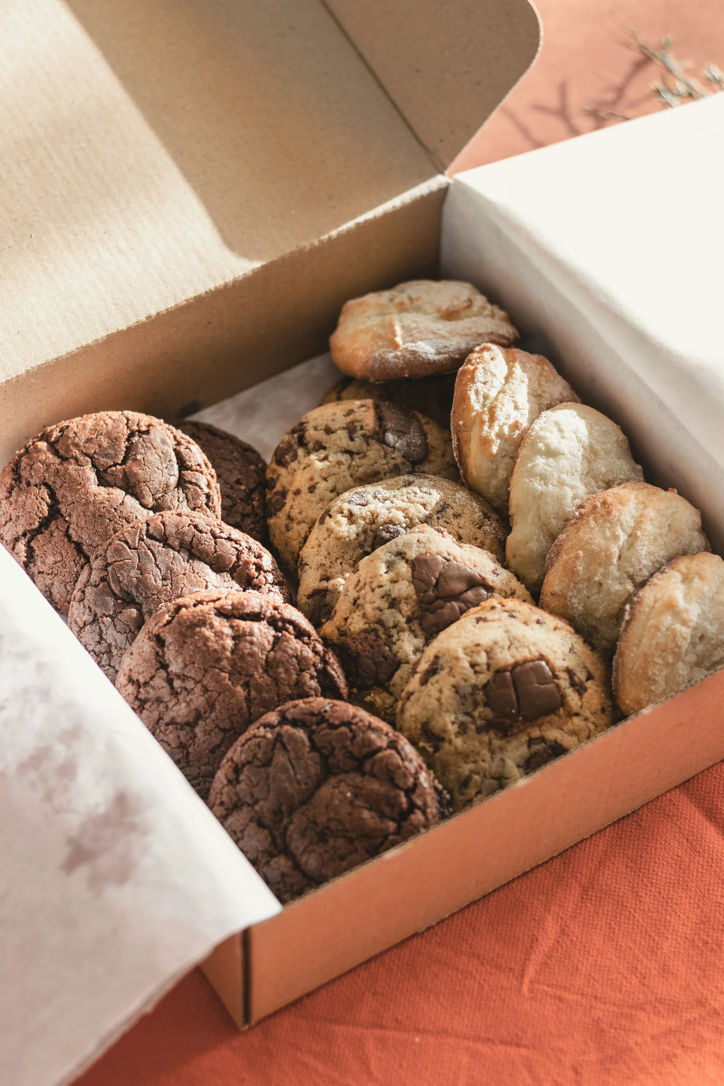 A box of assorted cookies, including chocolate chip, oatmeal, and double chocolate cookies on a wooden surface.