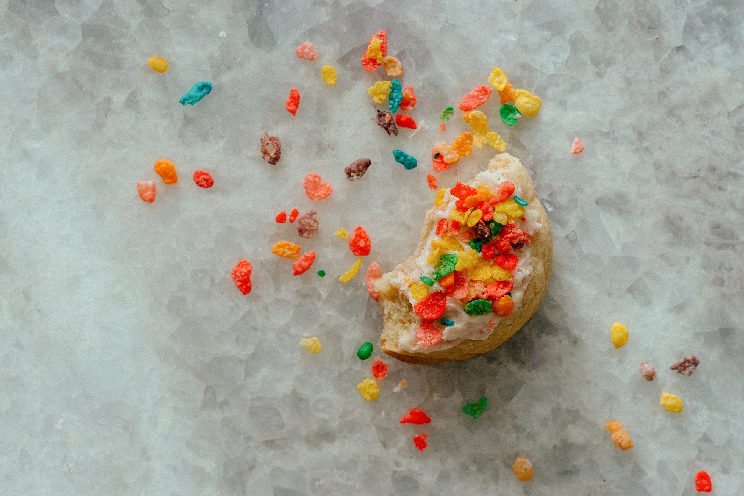 Half-eaten sugar cookie with white icing and colorful sprinkles, with scattered sprinkles on a marble surface.
