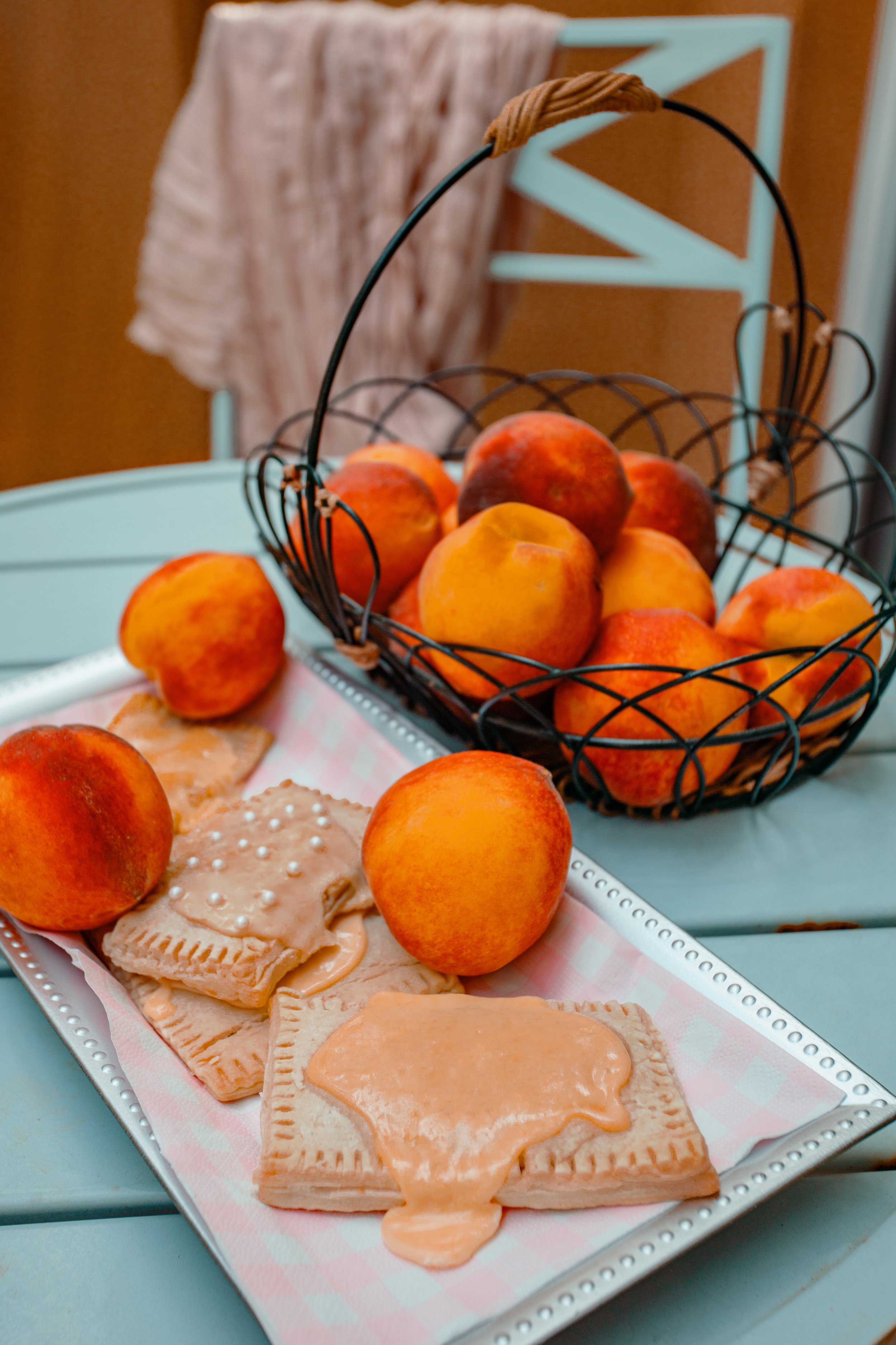 A tray of peach-flavored snacks with peach slices and peach-shaped candies, placed next to a black wire basket filled with fresh peaches, on a light blue table in an outdoor setting.