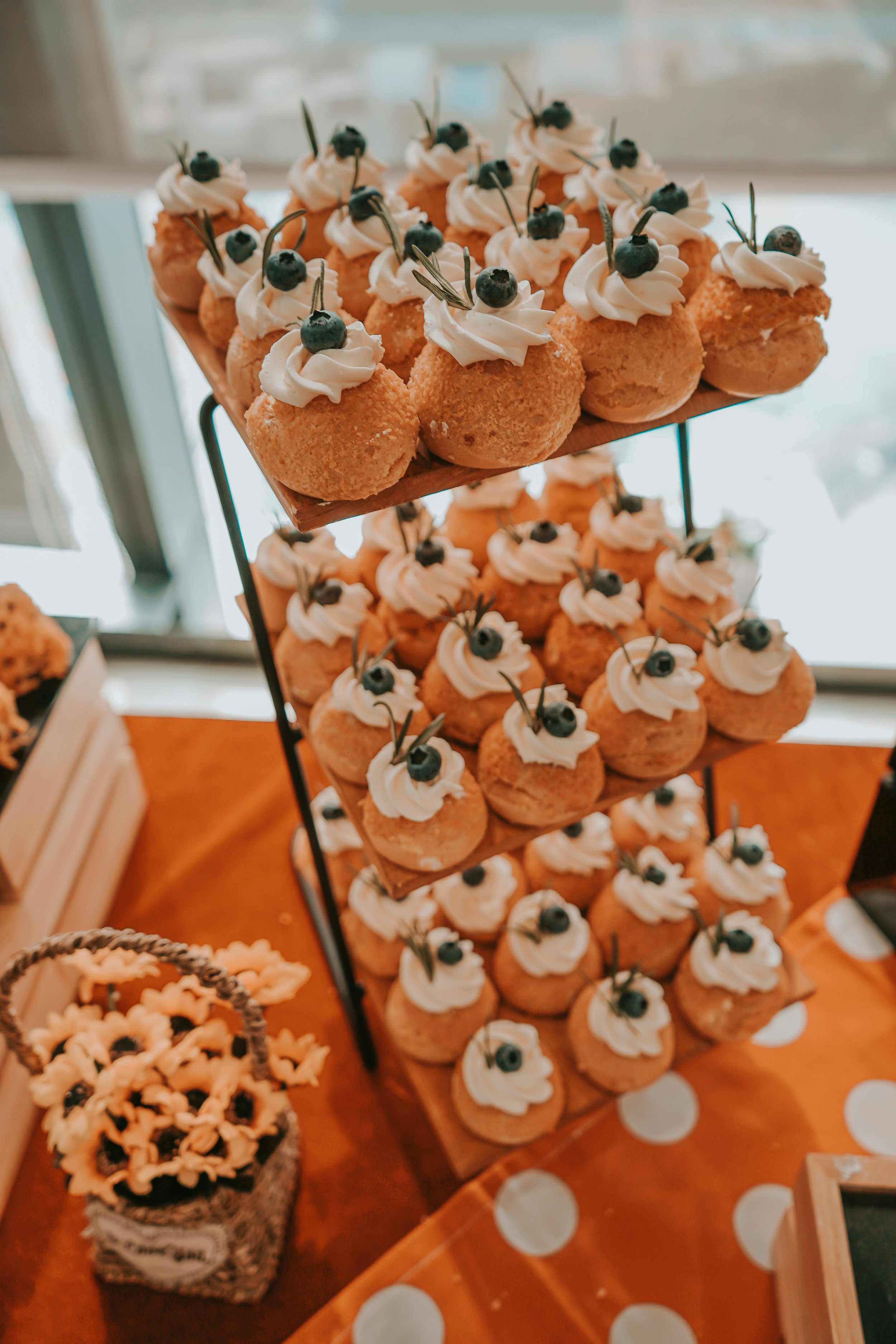 Display of mini cream puffs topped with whipped cream, blueberries, and decorative leaves on a metal stand, set on an orange tablecloth with white polka dots.