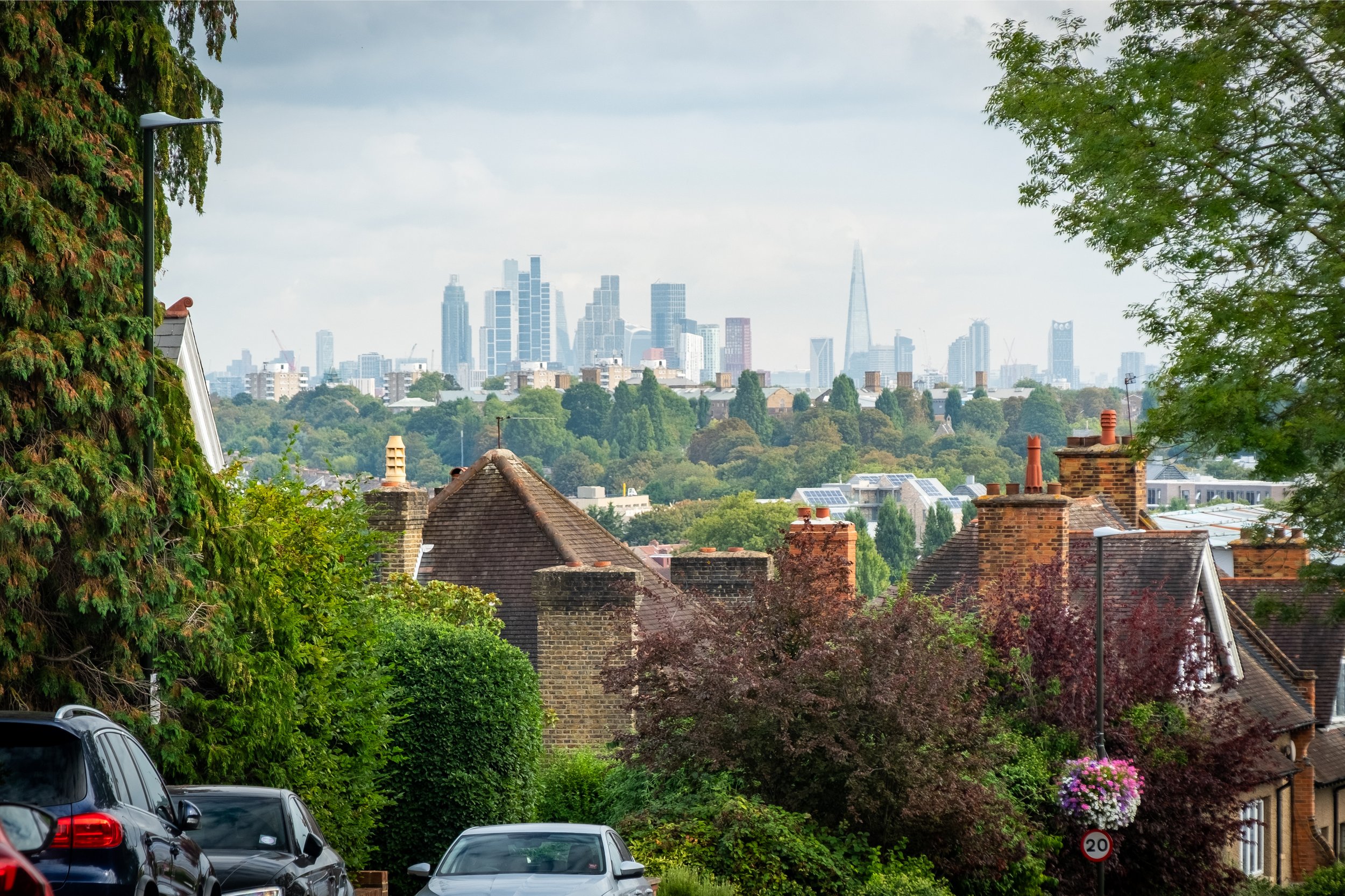 A view of a suburban neighborhood with cars parked along the street, surrounded by lush green trees, with the London skyline and The Shard in the background